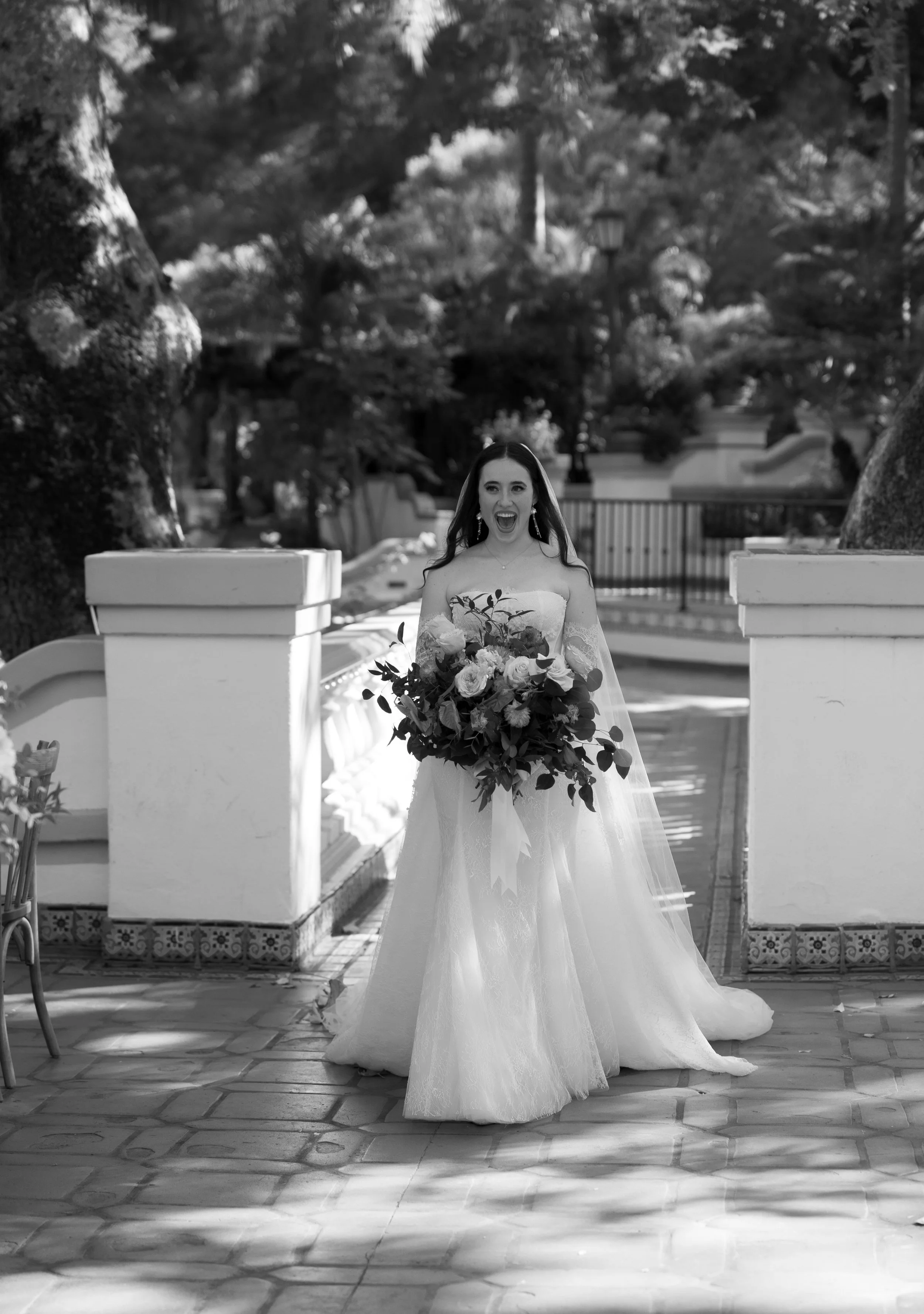 A bride in a wedding dress holding a large bouquet of flowers, standing outdoors on a paved area with trees and a fence in the background at a Rancho Las Lomas Wedding in Los Angeles
