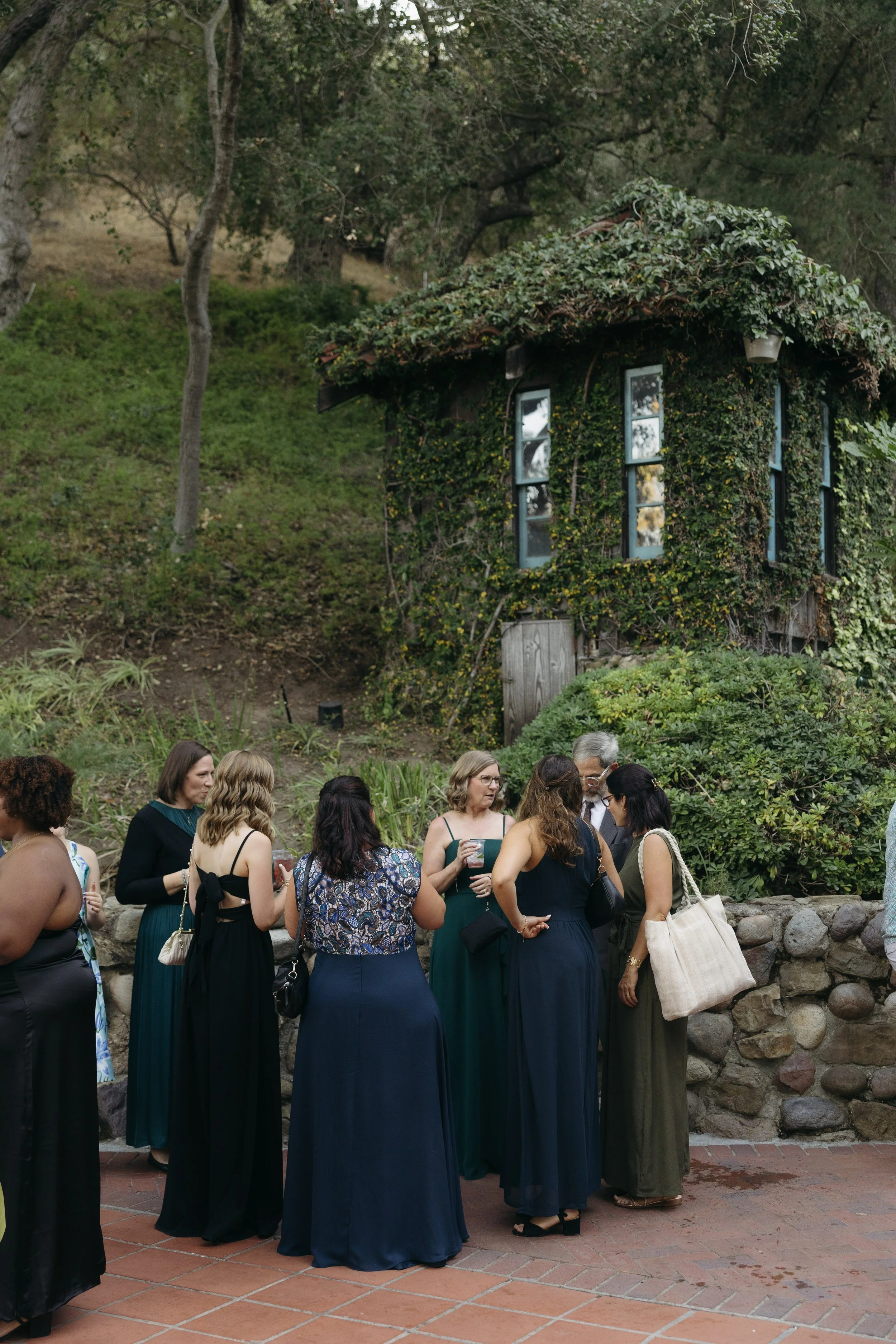 Group of women dressed in formal attire standing outdoors on a brick patio, engaged in conversation, with a small ivy-covered house on a grassy hillside in the background during cocktail hour at a Rancho Las Lomas Wedding in Los Angeles