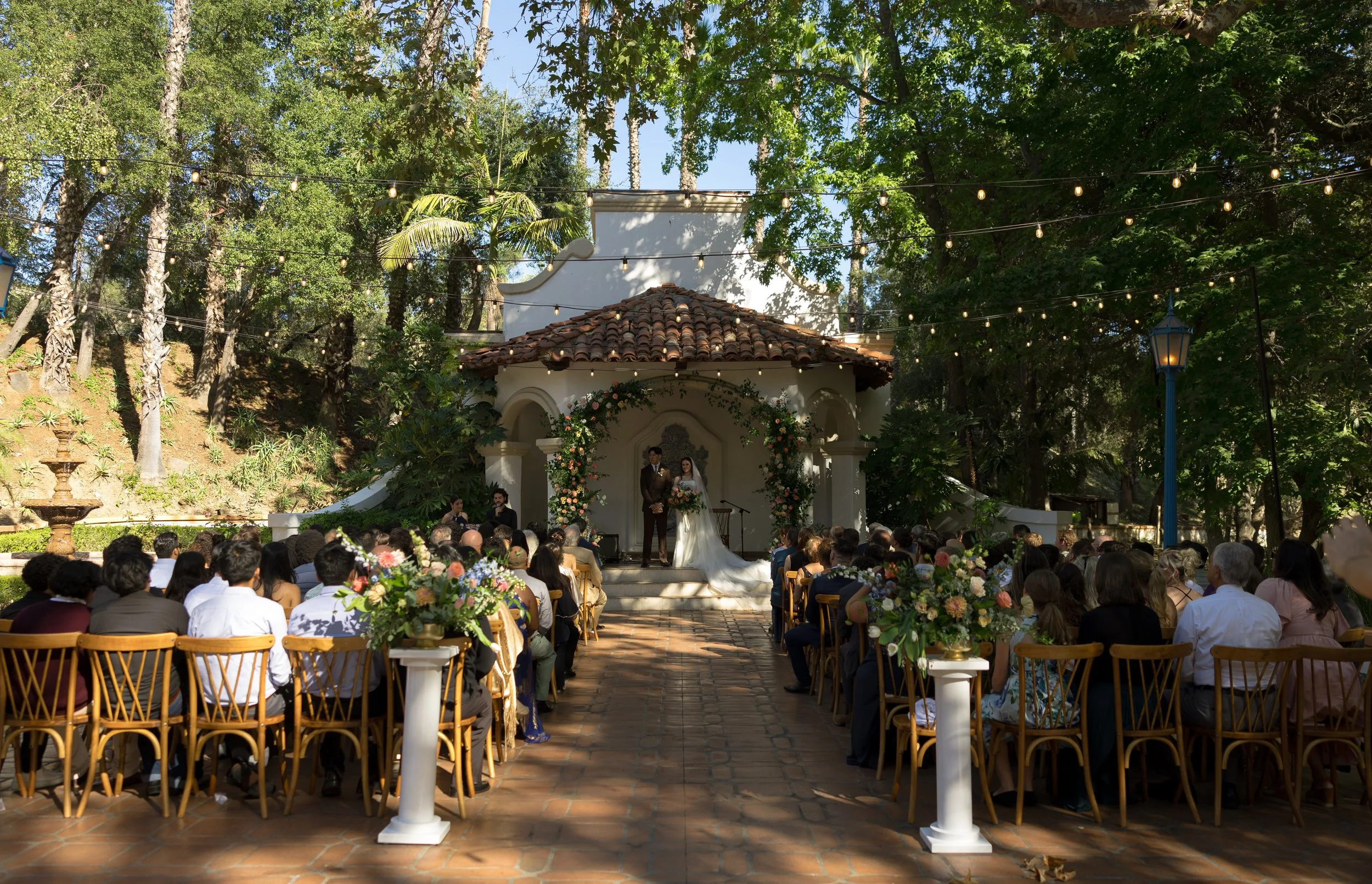 Outdoor wedding ceremony with bride and groom standing on a small stage under a floral arch, surrounded by seated guests, during daytime with trees and string lights overhead at a Rancho Las Lomas Wedding in Los Angeles