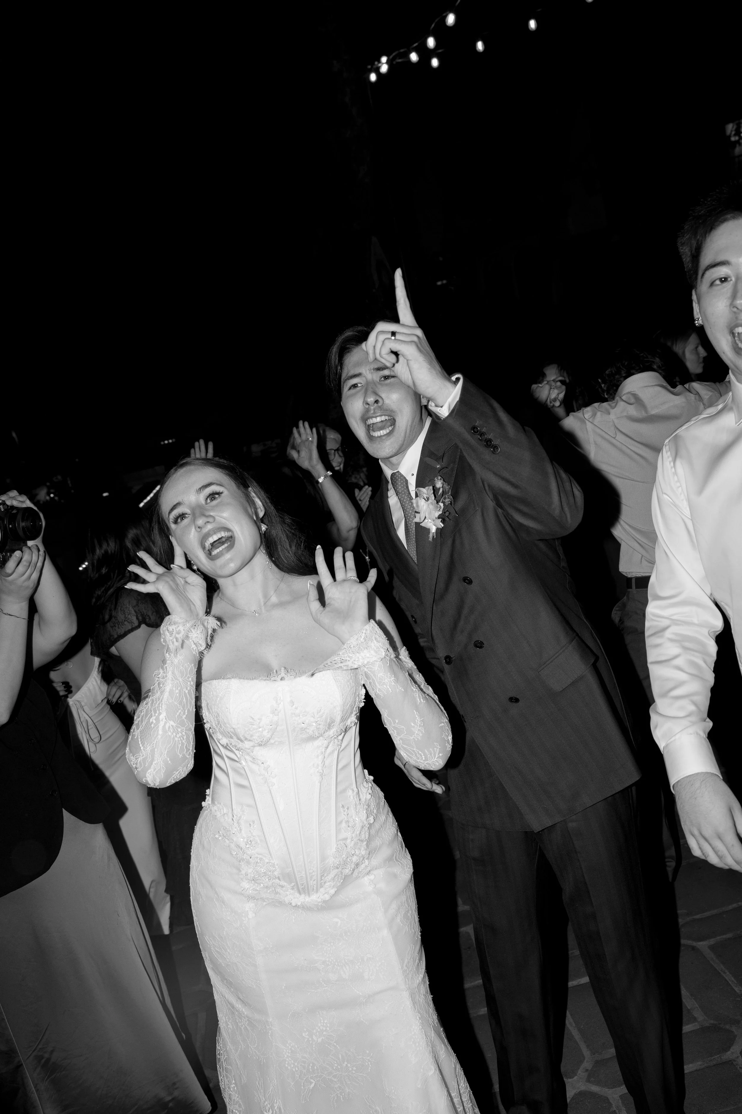 Black and white photo of a bride and groom celebrating at their wedding reception, with guests dancing around them.