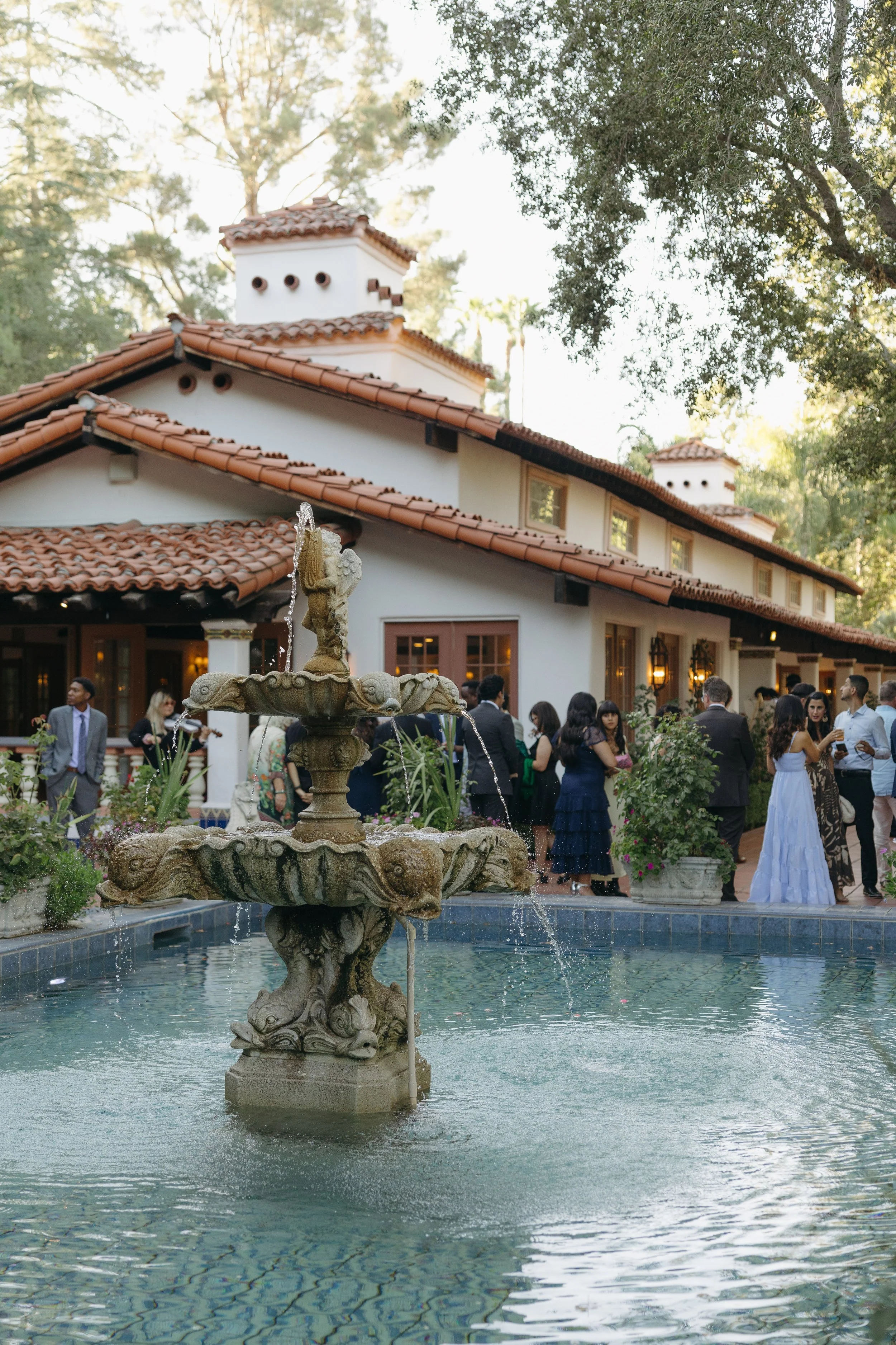 People attending a social event at a courtyard with a fountain, in front of a Spanish-style building with a tiled roof during cocktail hour at a Rancho Las Lomas Wedding in Los Angeles