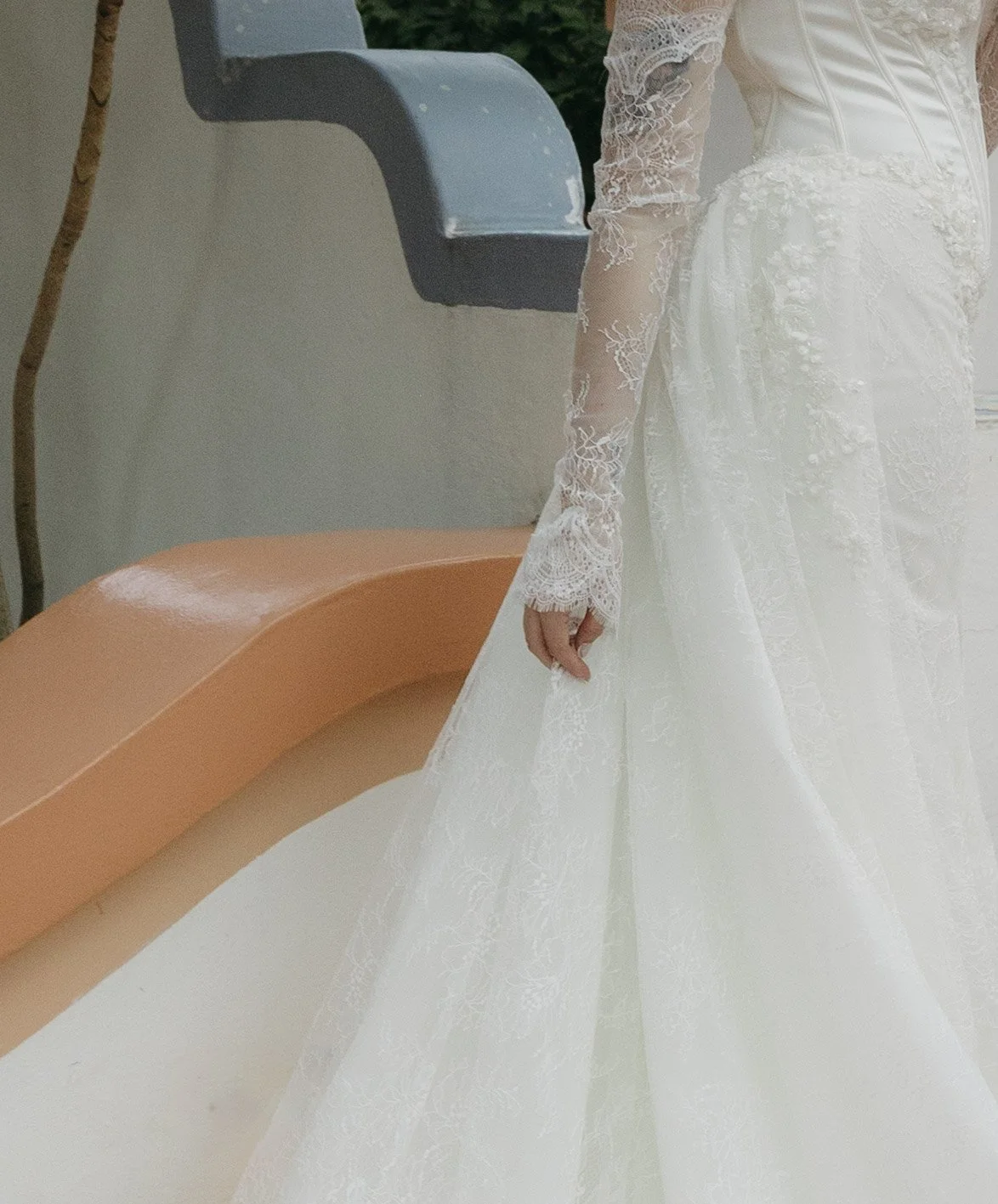 Part of a bride in a white lace wedding dress with long sleeves, standing next to a pastel orange bench with a grey backrest, holding the edge of her dress.