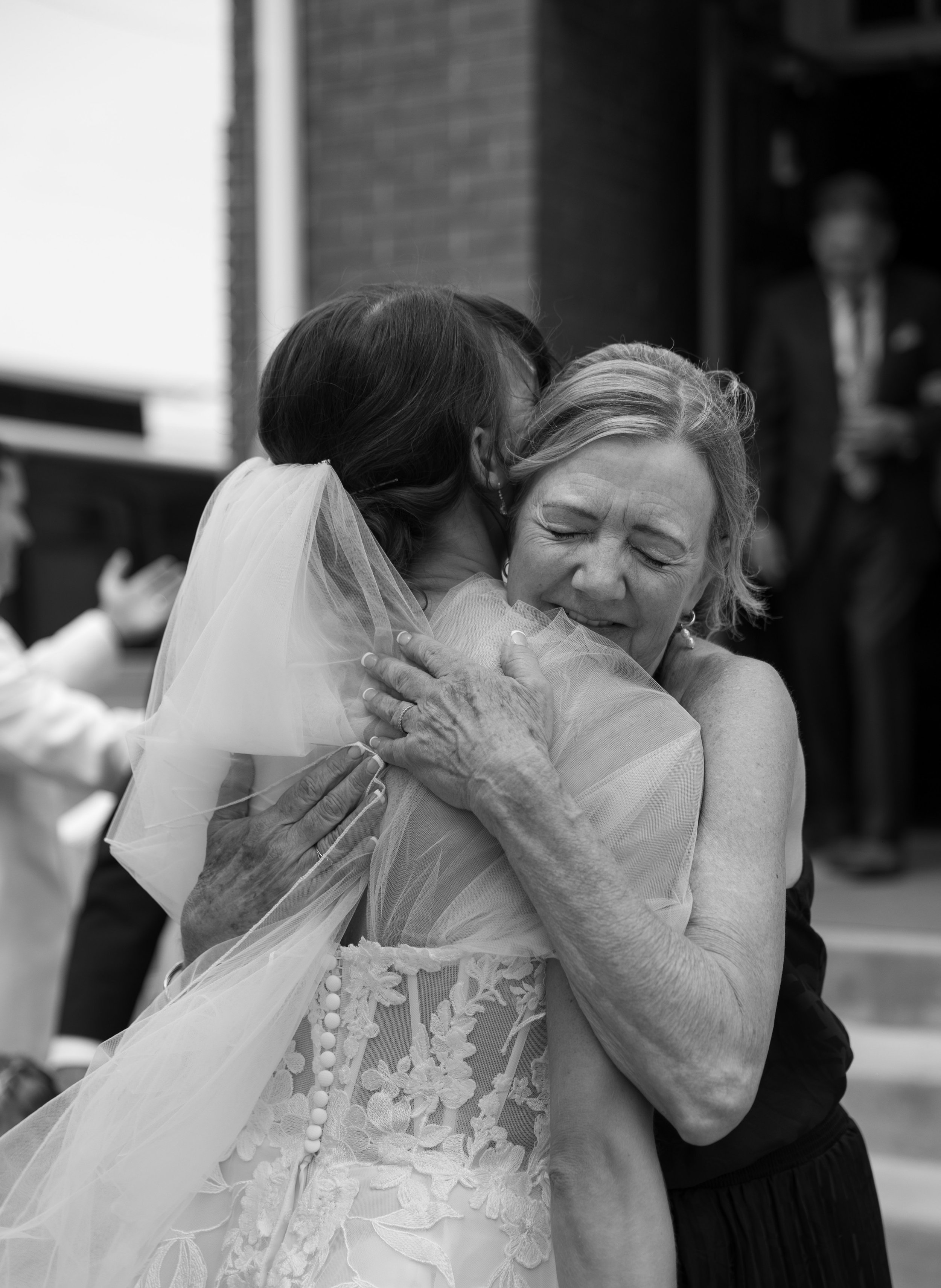 A woman in a wedding dress hugging her mother, who has her eyes closed and a contented smile, at a wedding celebration.