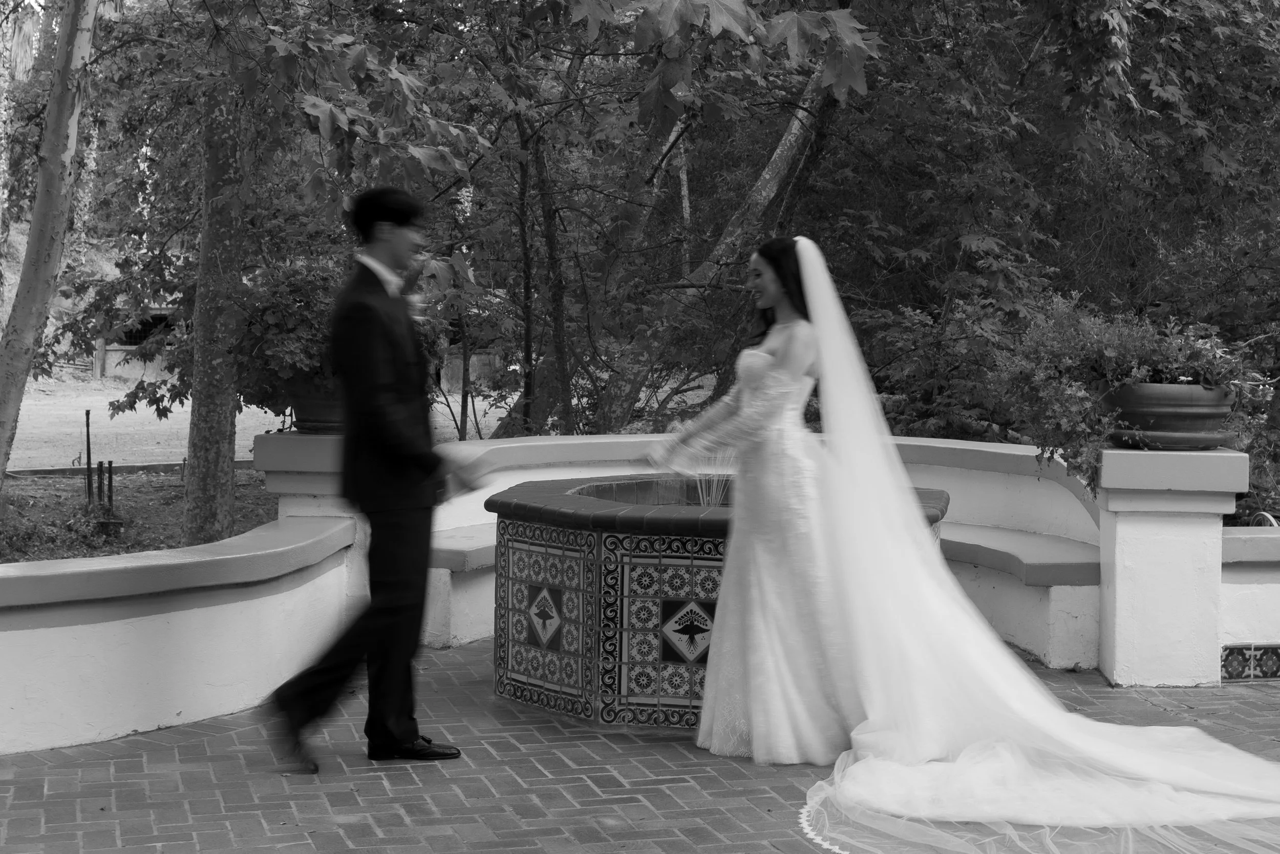 A bride and groom standing on a brick patio with a decorative water fountain, surrounded by trees and potted plants.