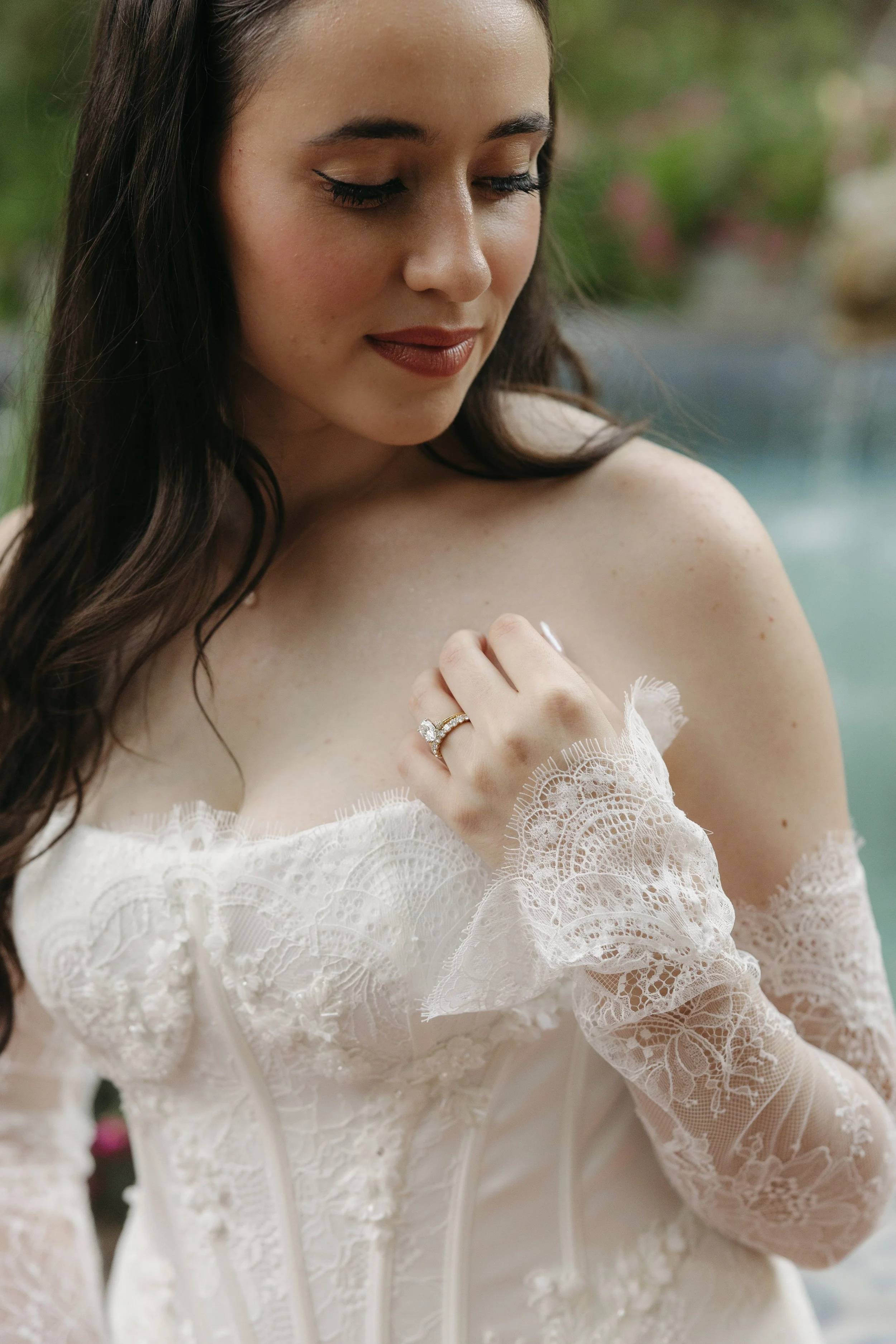 Close-up of a woman in an off-the-shoulder lace dress with long lace gloves, wearing a wedding ring, with her eyes closed and a serene expression, outdoors near a pool.