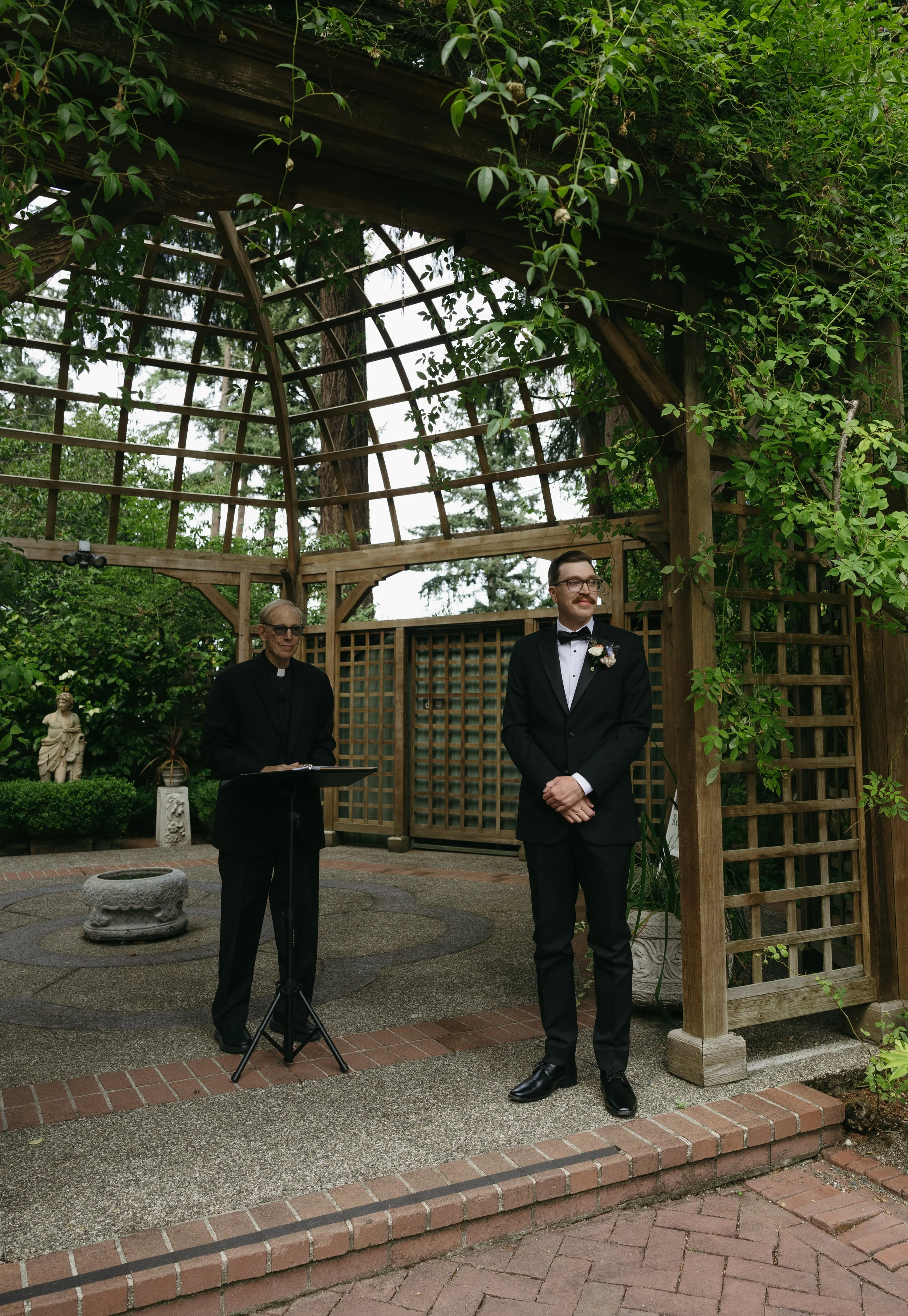 A wedding ceremony taking place outdoors in a garden with a wooden arbor decorated with green vines. A man in a tuxedo with a boutonniere stands smiling while a clergyman in black robes stands nearby with a book.