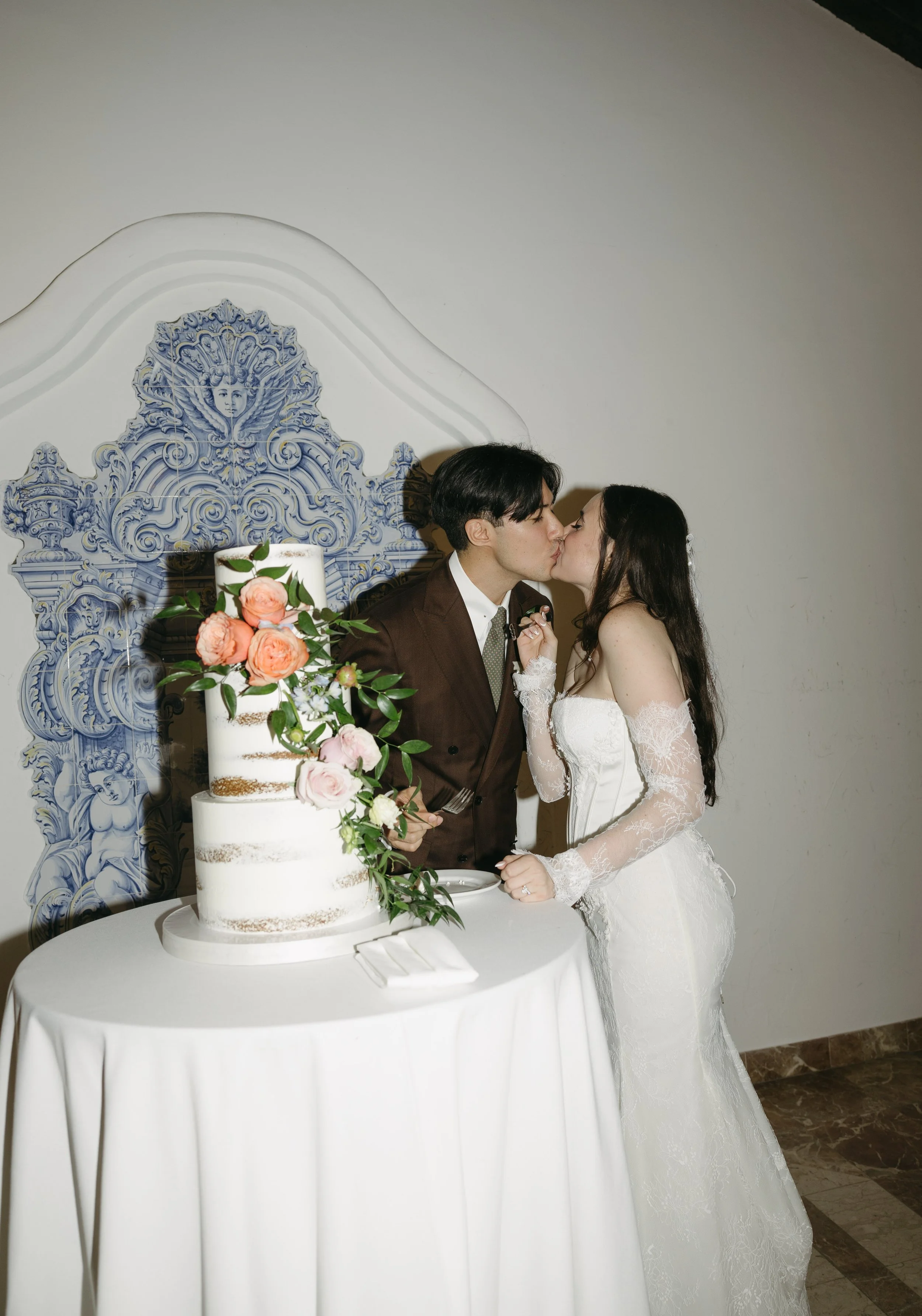 A bride and groom kissing during their wedding reception, standing next to a tiered wedding cake decorated with pink and peach flowers and green foliage.