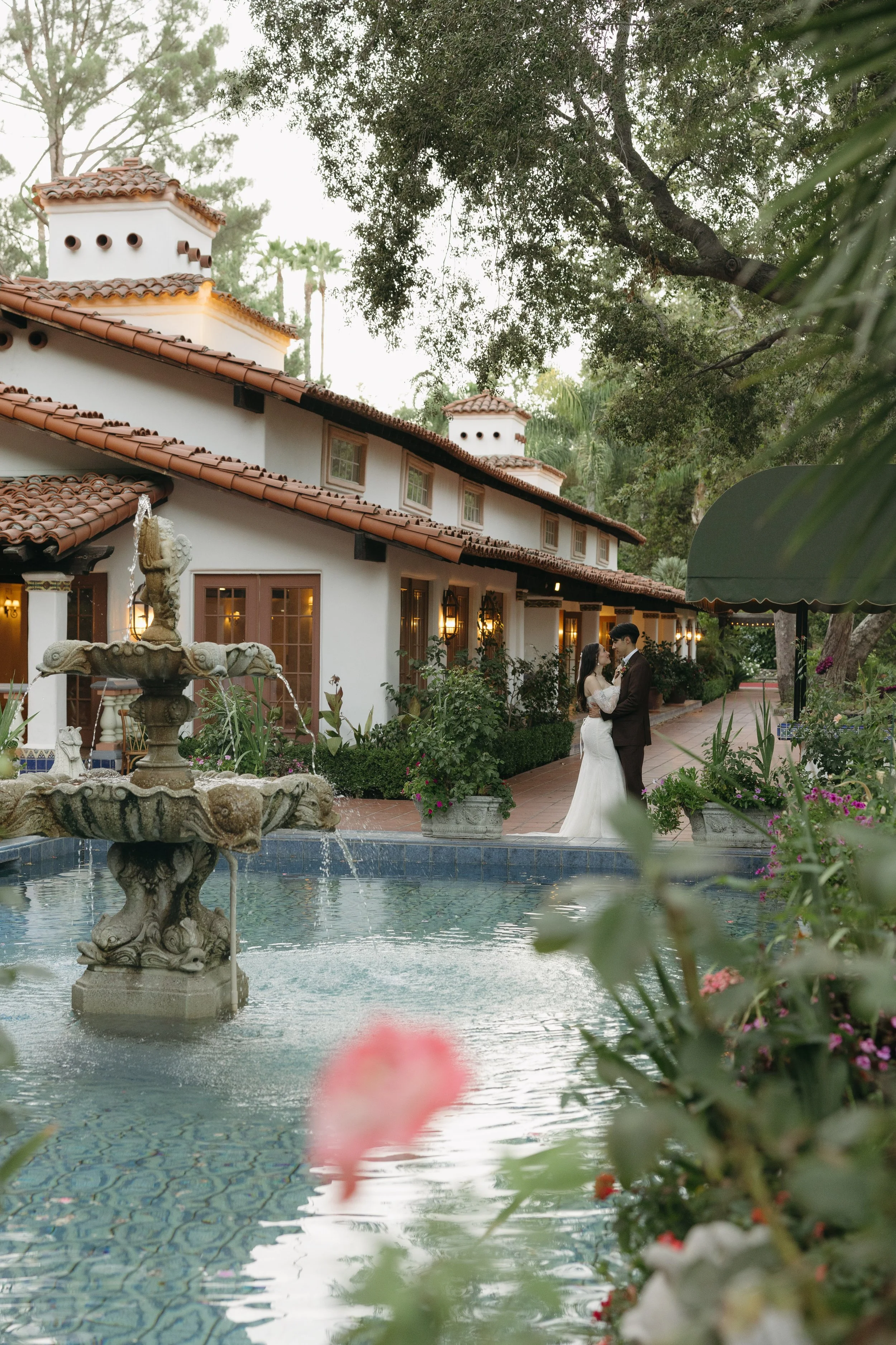 A bride and groom in wedding attire embracing near a fountain outside a Spanish-style building with white stucco walls, red tile roof, and palm trees in the background at a Rancho Las Lomas Wedding in Los Angeles