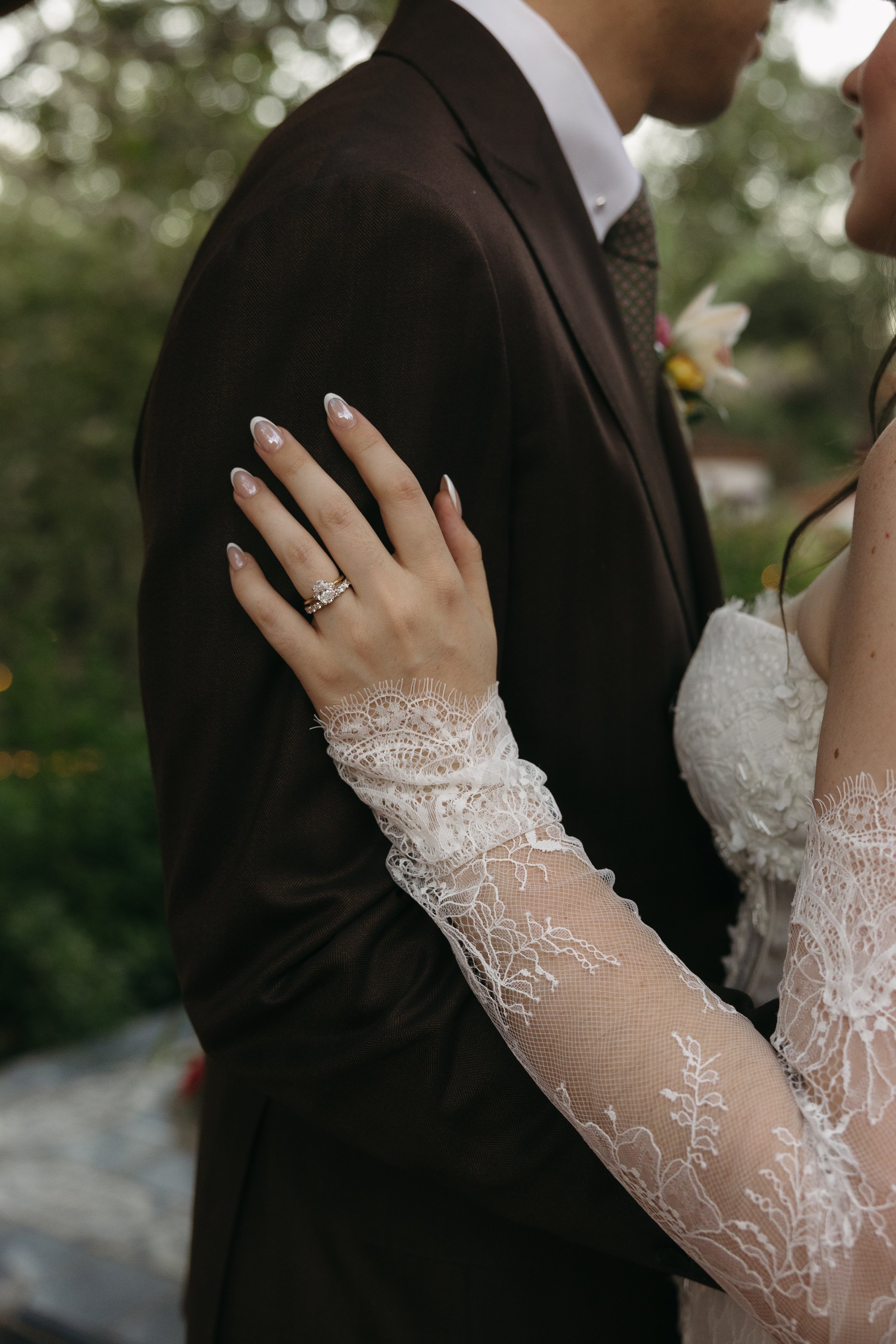 Close-up of a wedding couple, focusing on the bride's hand with a wedding ring on her finger, resting on the groom's chest. The bride is wearing lace sleeves, and the groom is in a black suit with a white shirt and a bow tie. Outdoors with blurred gr