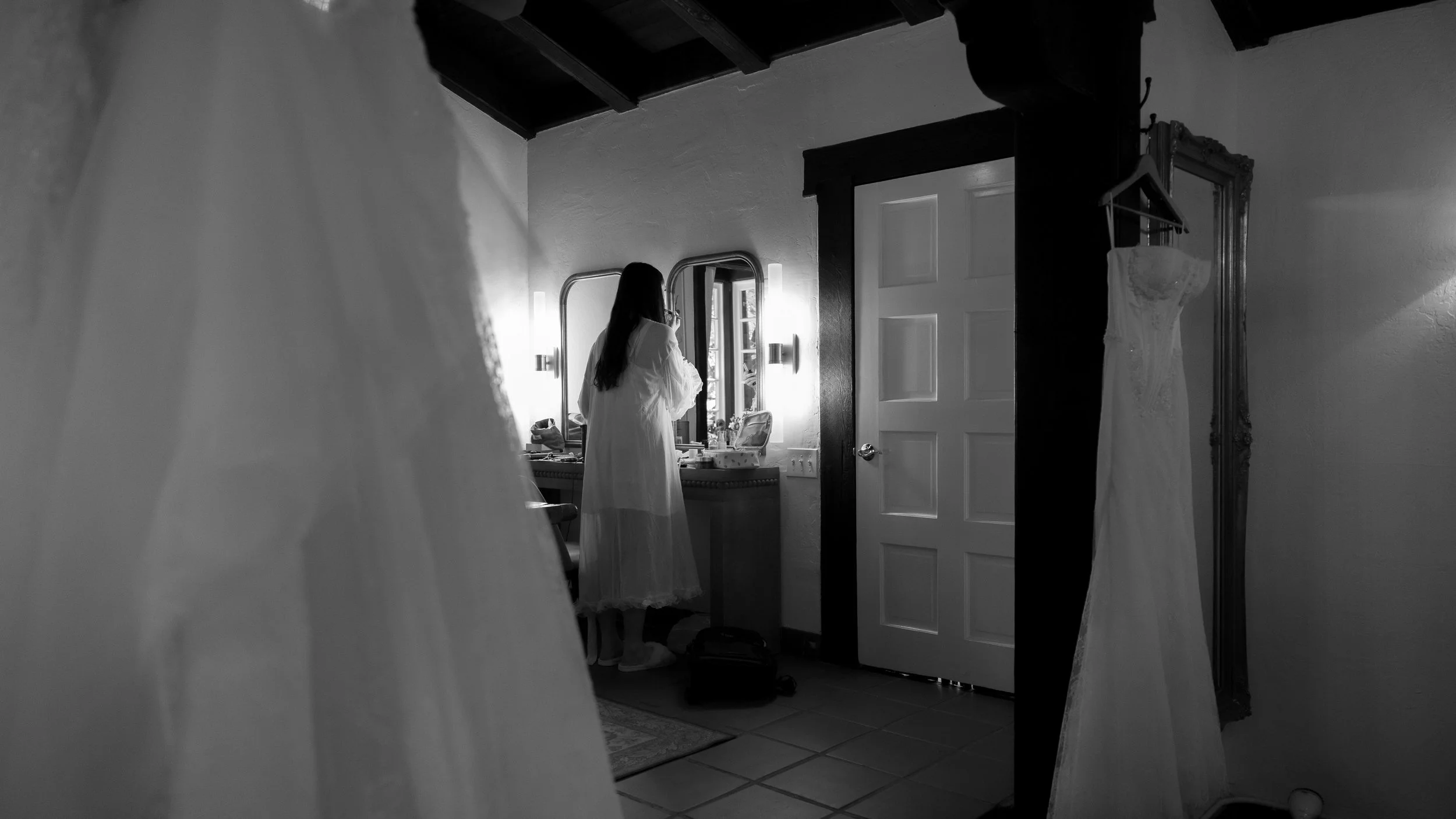A bride in a white dress is looking at herself in a mirror in a bedroom or dressing area, with a wedding dress hanging on the wall to the right at a Rancho Las Lomas Wedding in Los Angeles