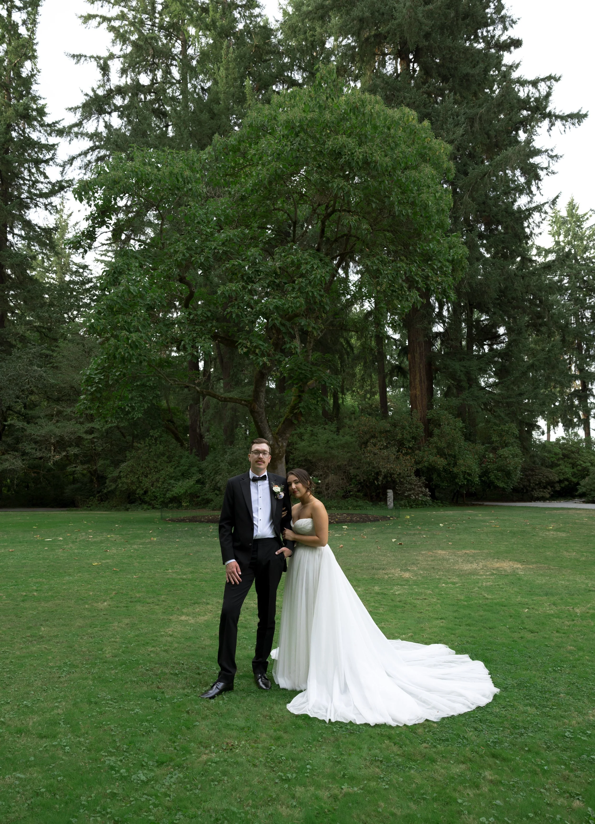 A bride and groom standing together on a grassy area in a park with large trees in the background at a European inspired Lakewold Gardens wedding in Seattle