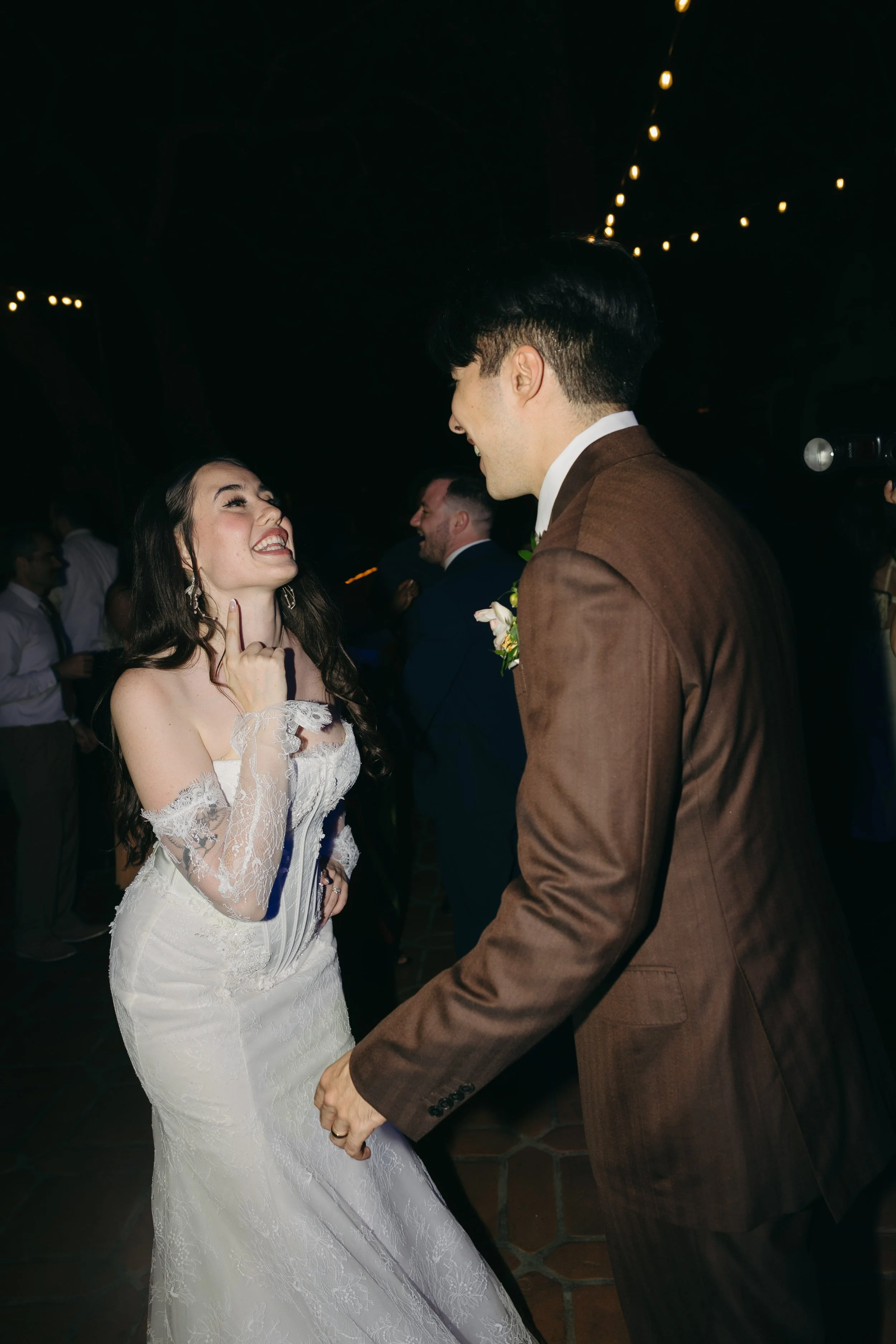 A bride and groom dancing and smiling at their wedding reception, with other guests in the background and string lights above them.