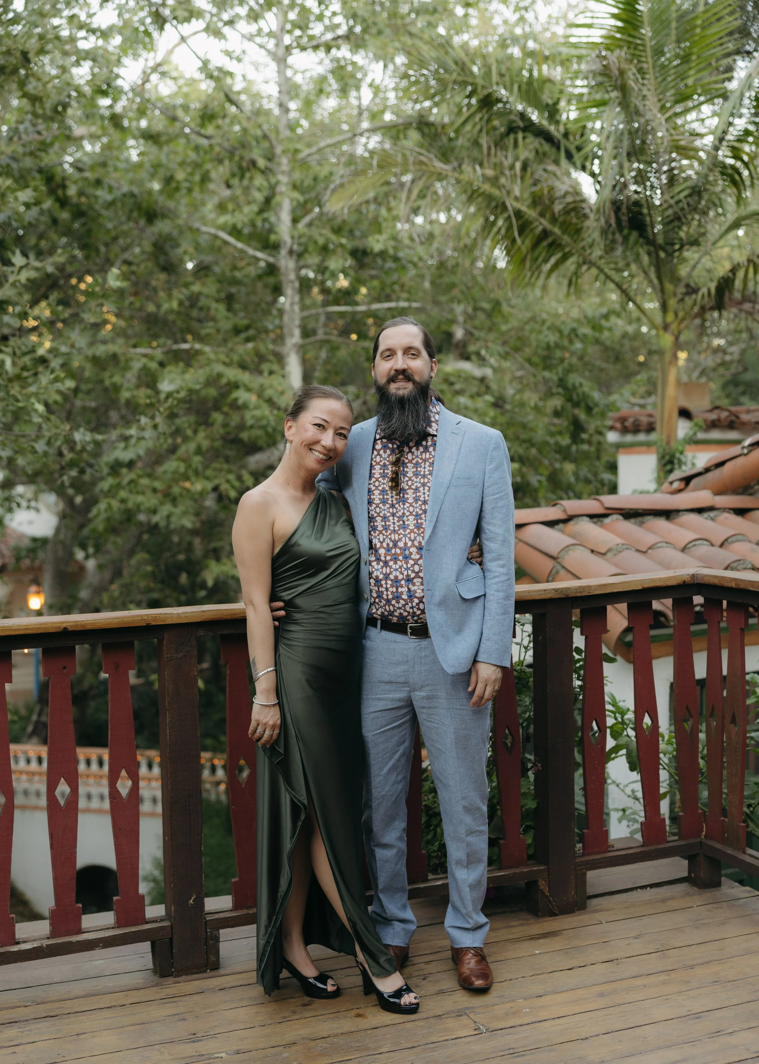 A couple standing on a wooden deck outdoors, with trees and a tiled roof in the background. The woman is wearing a dark green, one-shoulder gown and the man is dressed in a light blue suit with a patterned shirt. They are smiling and posing together.