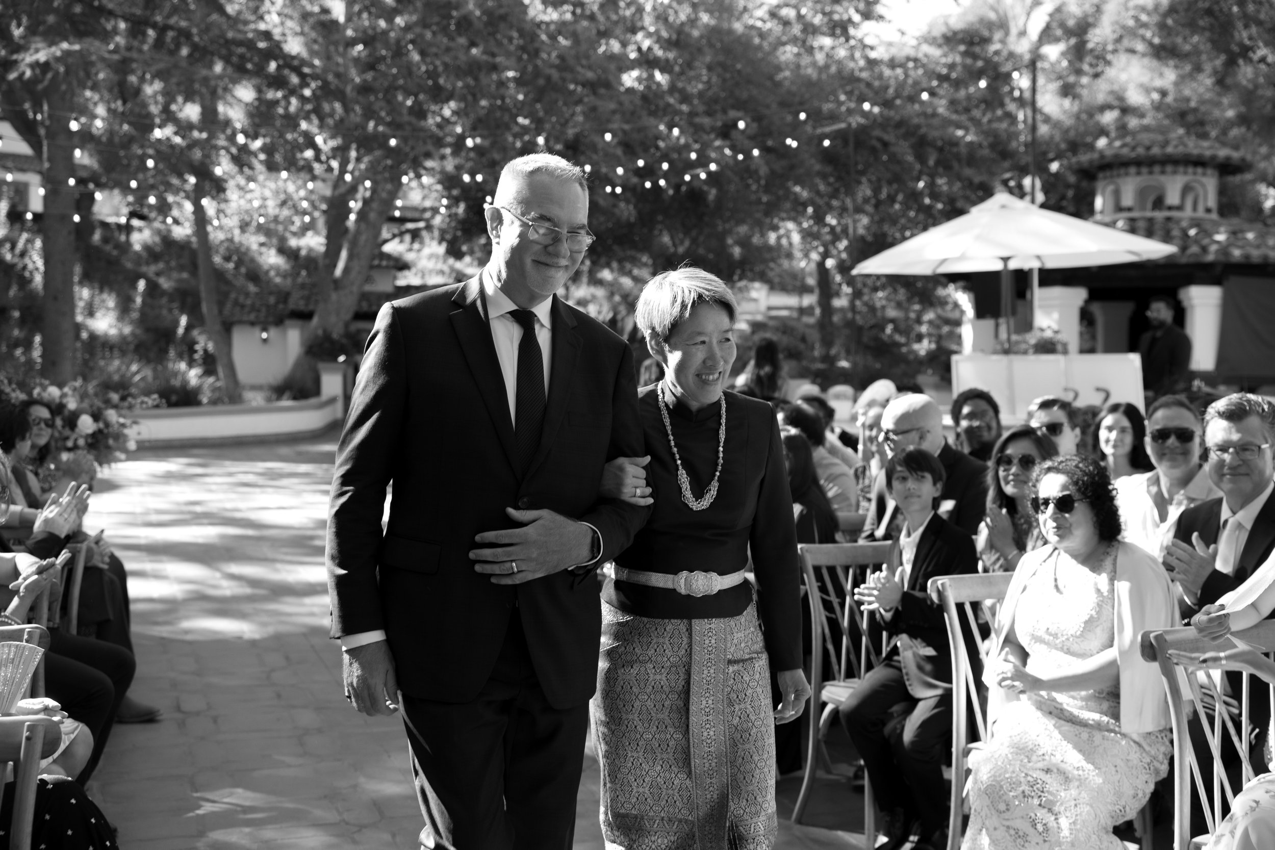 A black-and-white photo of a man in a suit walking down an aisle with a woman in a black outfit and patterned skirt, surrounded by seated guests at an outdoor event, possibly a wedding, with trees and string lights in the background.