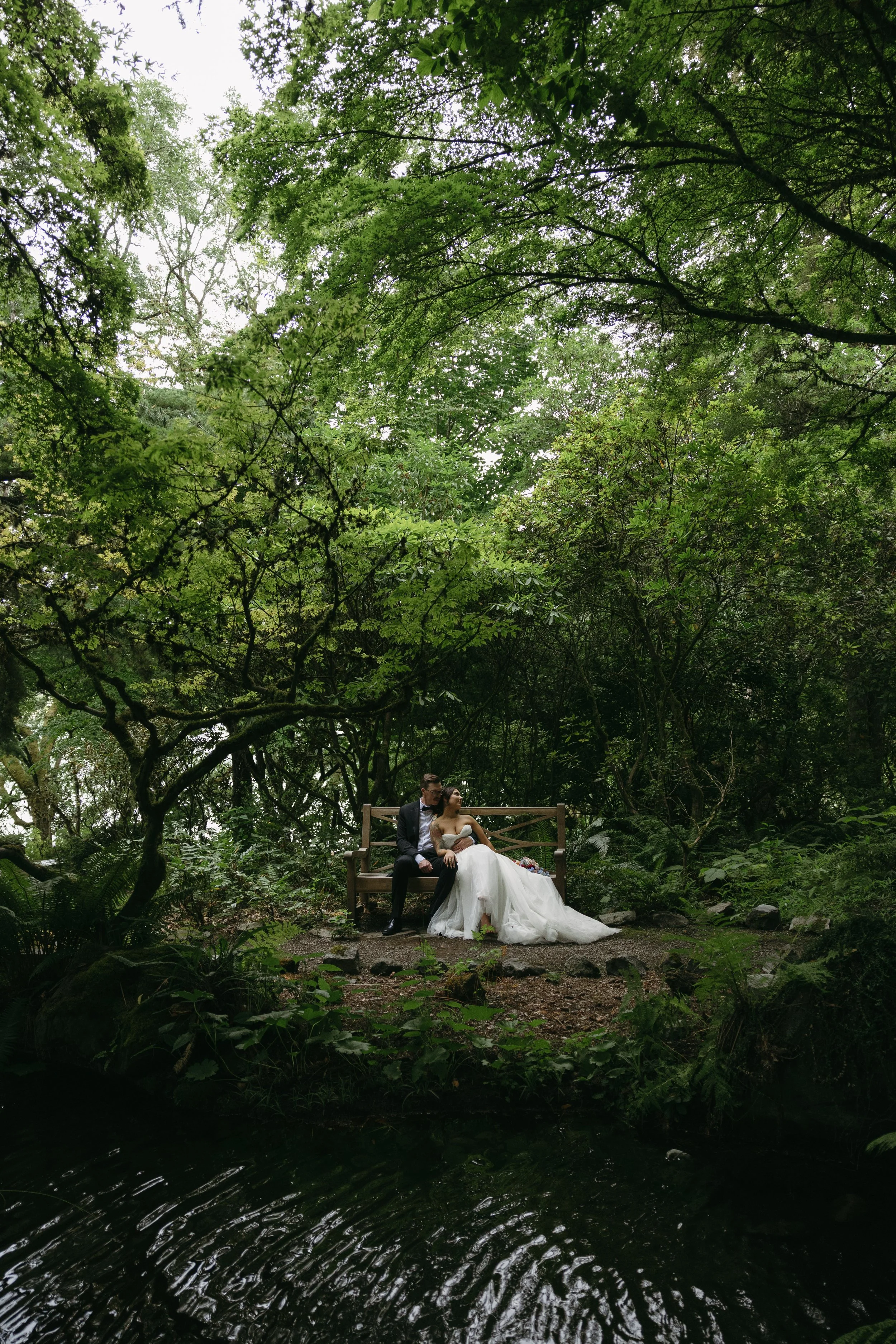 A couple in wedding attire sitting on a wooden bench in a lush, green forest near a small body of water at a European inspired Lakewold Gardens wedding in Seattle