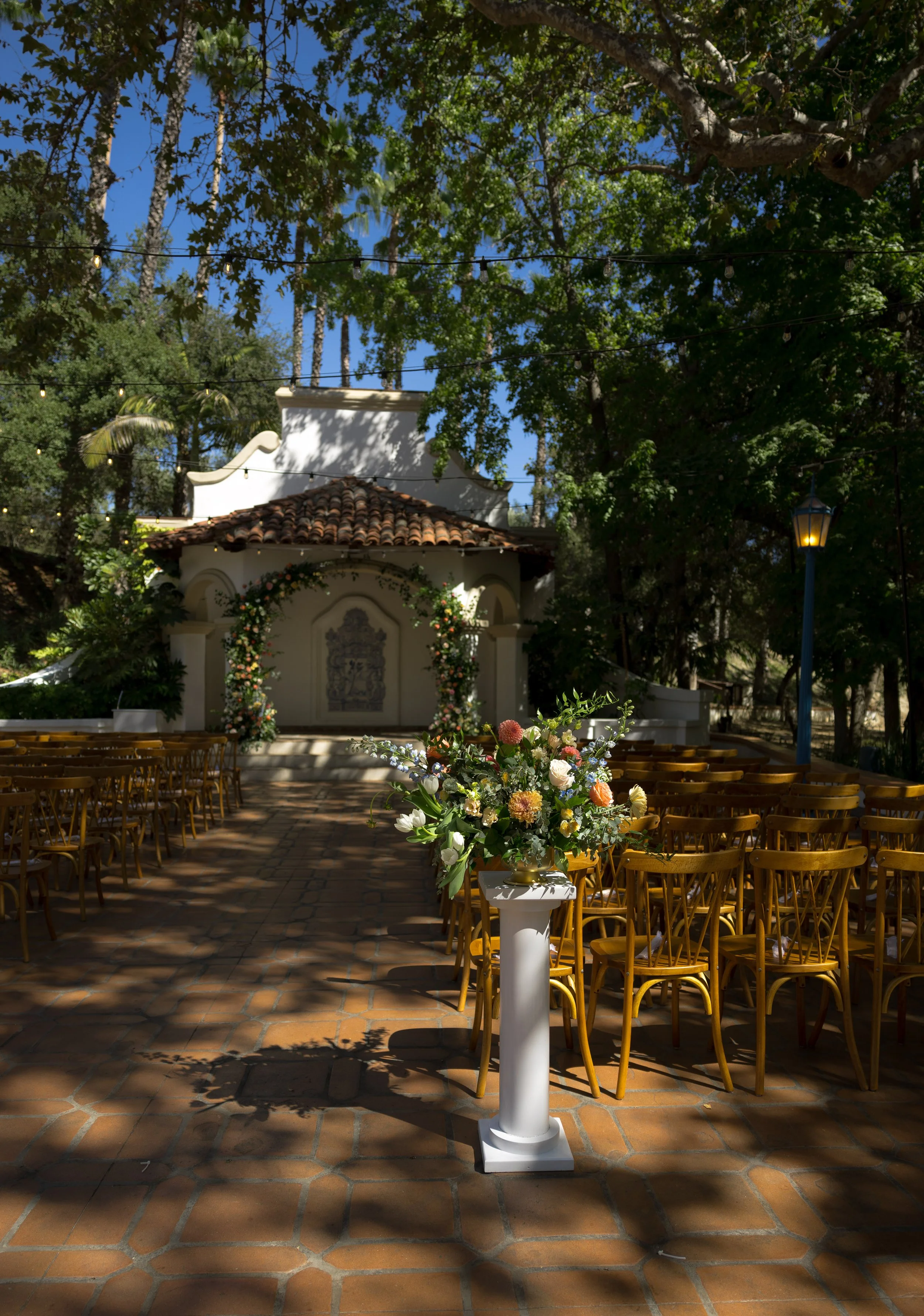 Outdoor wedding ceremony setup with rows of wooden chairs on brick paving, a floral arrangement on a white pedestal, and a small chapel decorated with flowers in a lush, shaded garden at a Rancho Las Lomas Wedding in Los Angeles