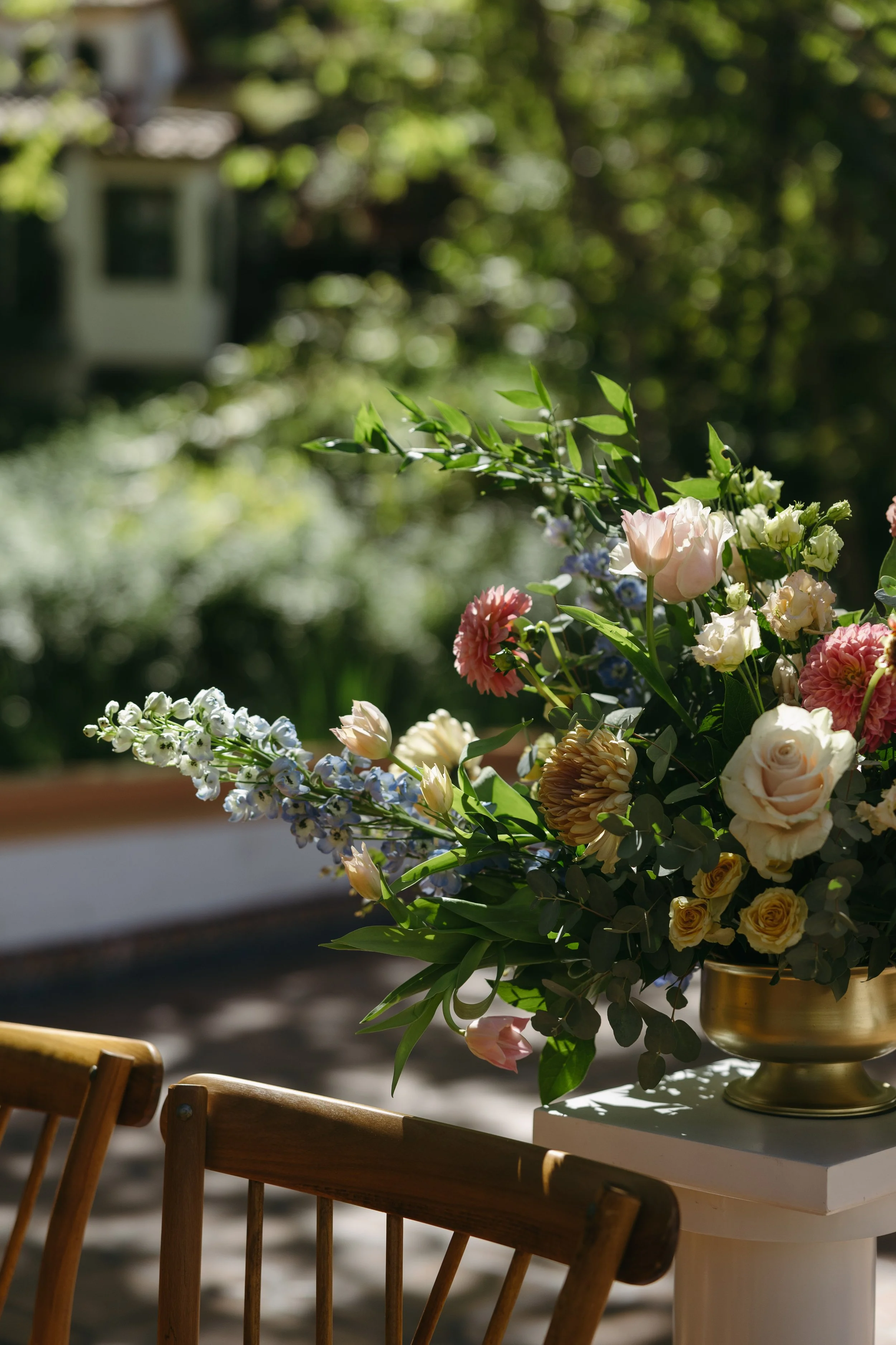 A large bouquet of assorted flowers including roses, tulips, and other blooms in a gold vase placed on a white table outdoors. Wooden chairs are nearby, and the background features a garden with lush greenery and trees.