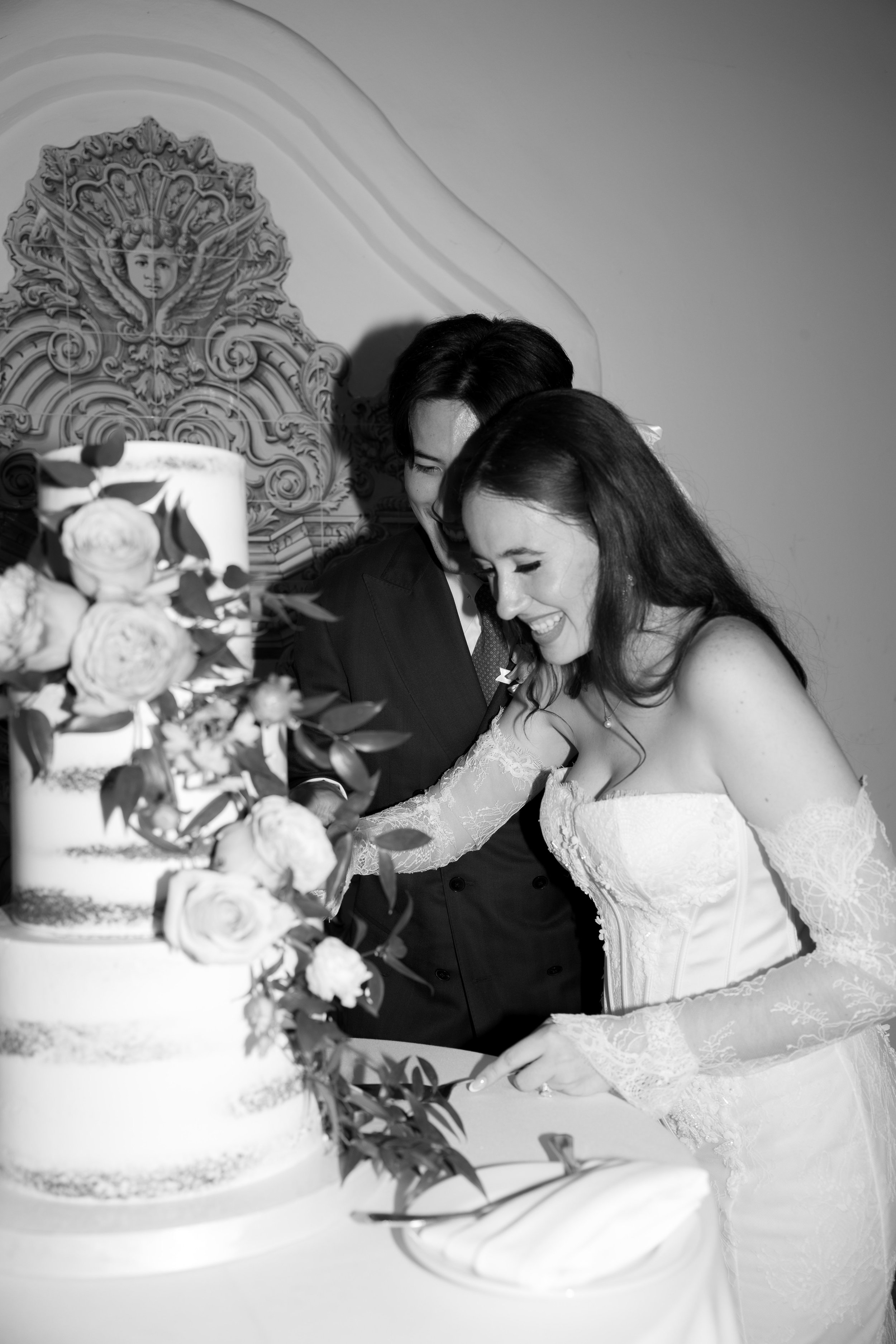 A bride and groom cutting a wedding cake together, smiling, with floral decorations on the cake and a decorative wall in the background.