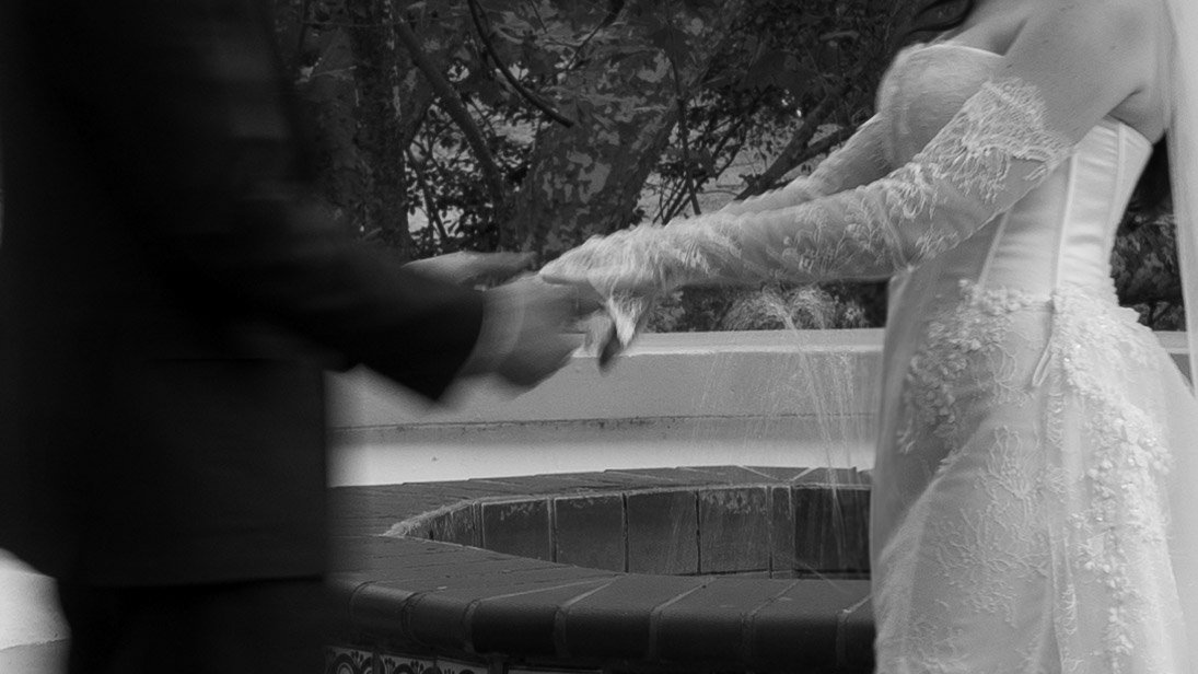 A couple holding hands during their wedding ceremony, with the bride wearing a lace wedding dress and the groom in a dark suit, outdoors near a fountain.