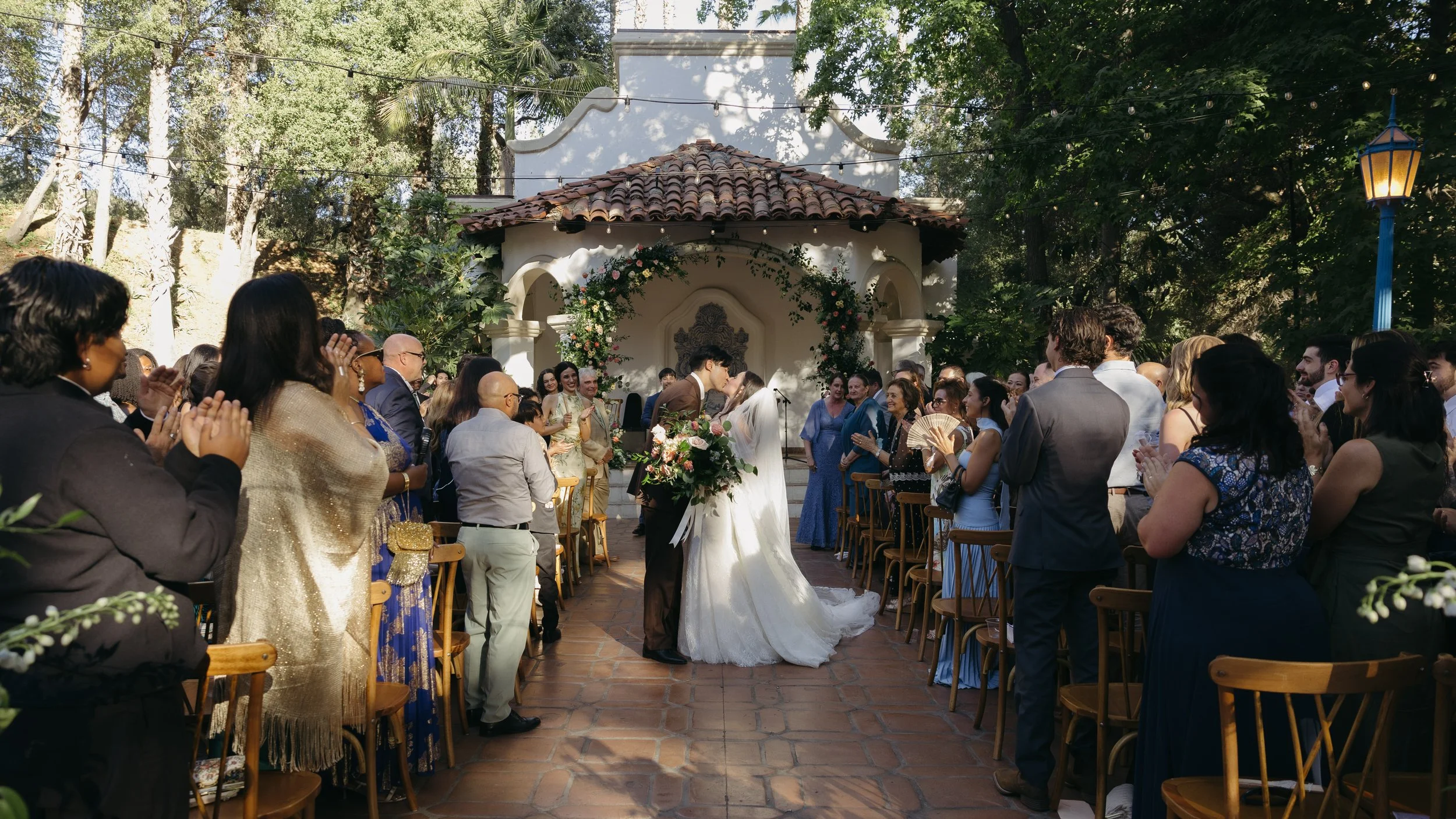 Outdoor wedding ceremony with a bride and groom kissing, surrounded by standing guests clapping, in a garden with trees, greenery, and a decorative arch and floral decor in the background at a Rancho Las Lomas Wedding in Los Angeles