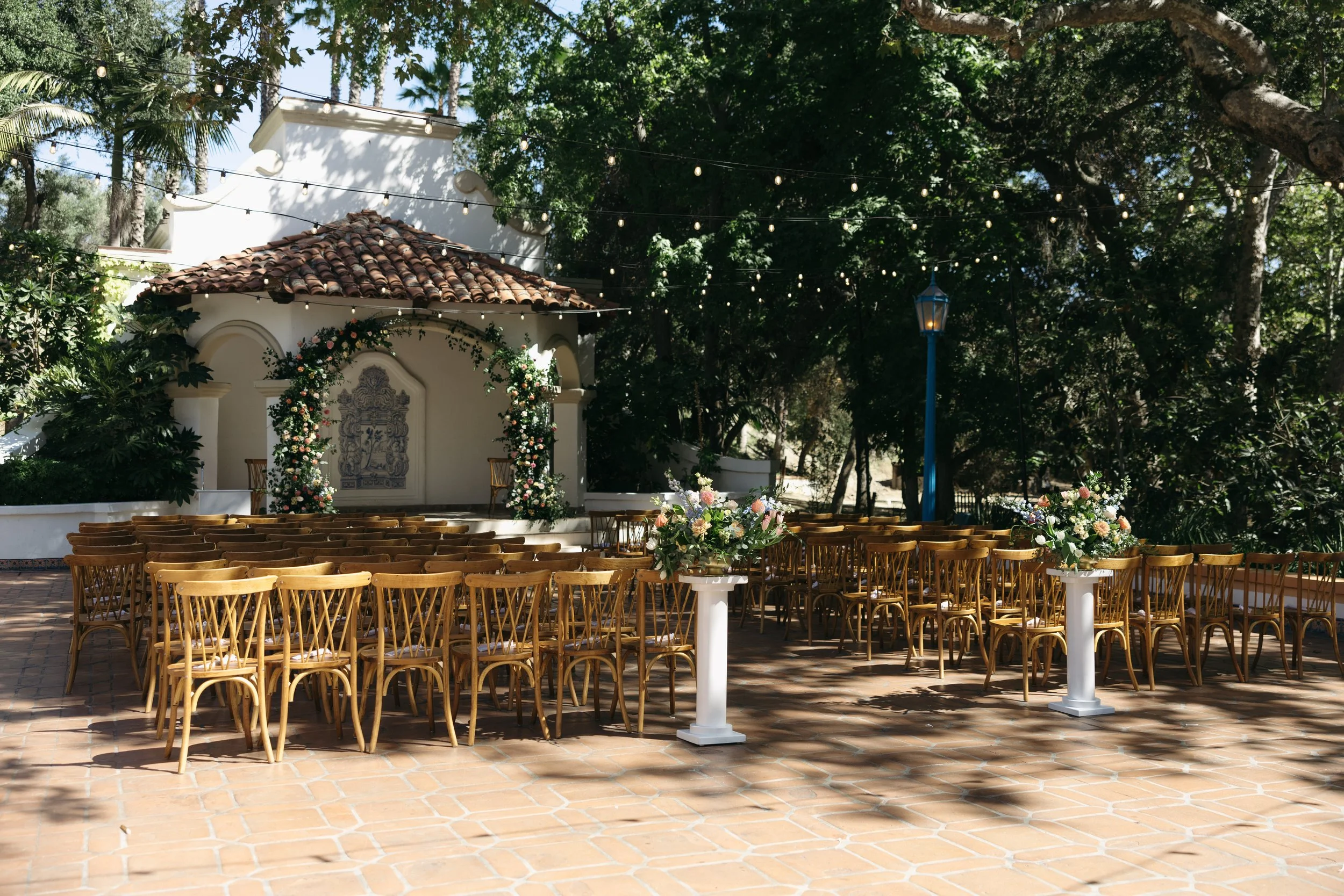 Outdoor wedding ceremony setup with rows of wooden chairs, floral arrangements, a small stage with a decorative wall, and string lights surrounded by trees at a Rancho Las Lomas Wedding in Los Angeles