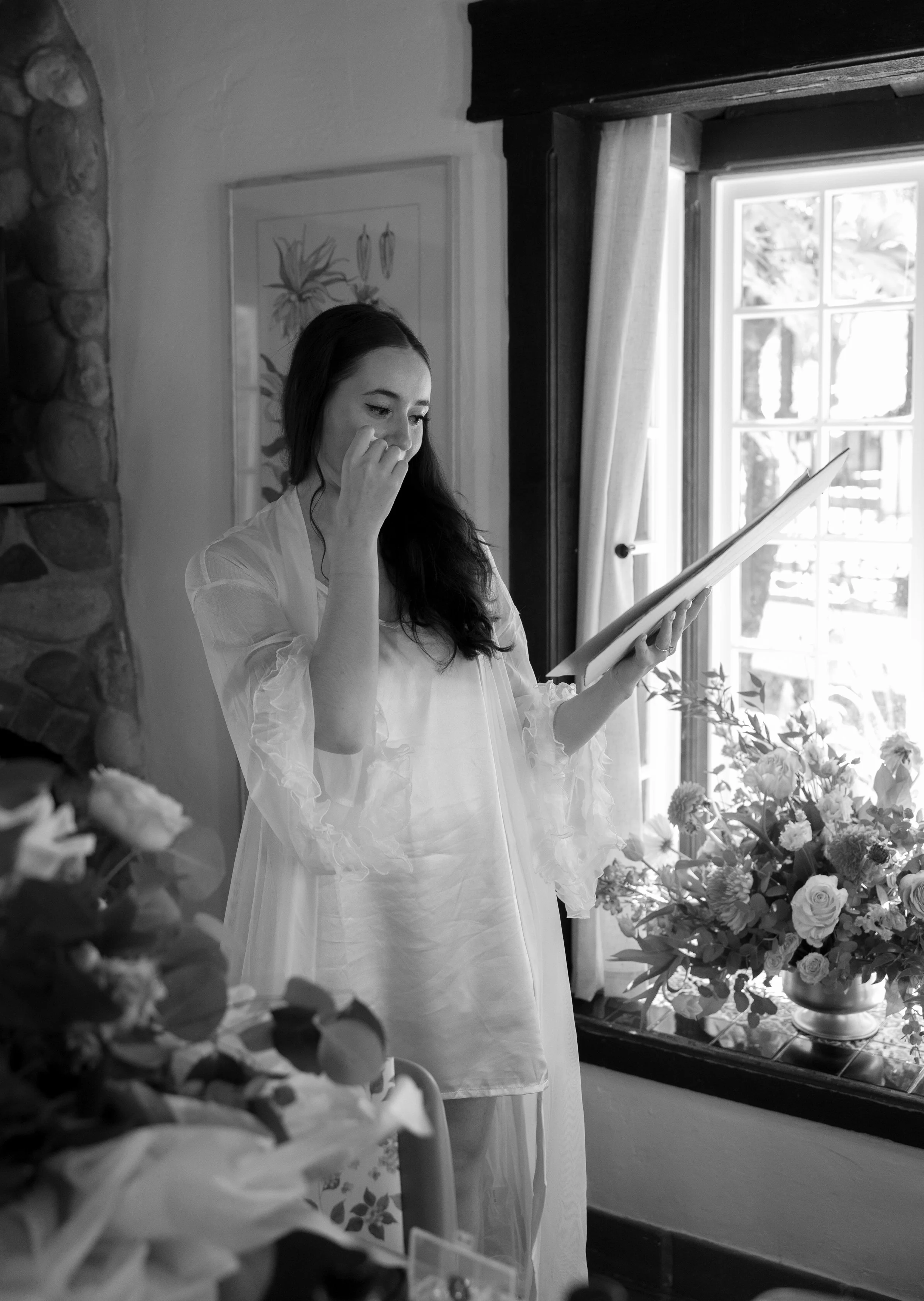 A woman in a white dress reading a book or card near a window with flowers on a table.
