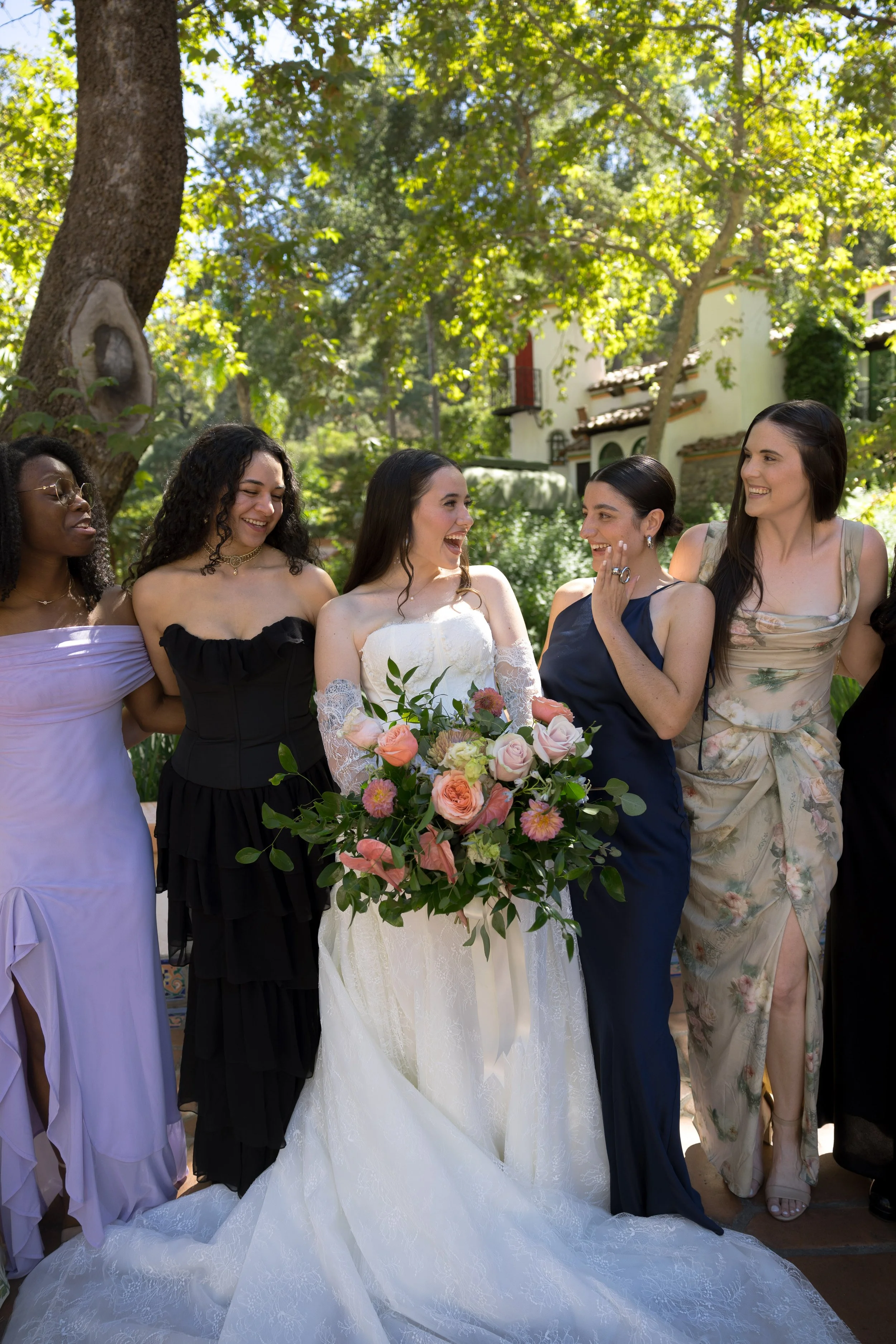 A group of bridesmaids, including a bride in a white wedding dress holding a bouquet, celebrating outdoors among trees and greenery.