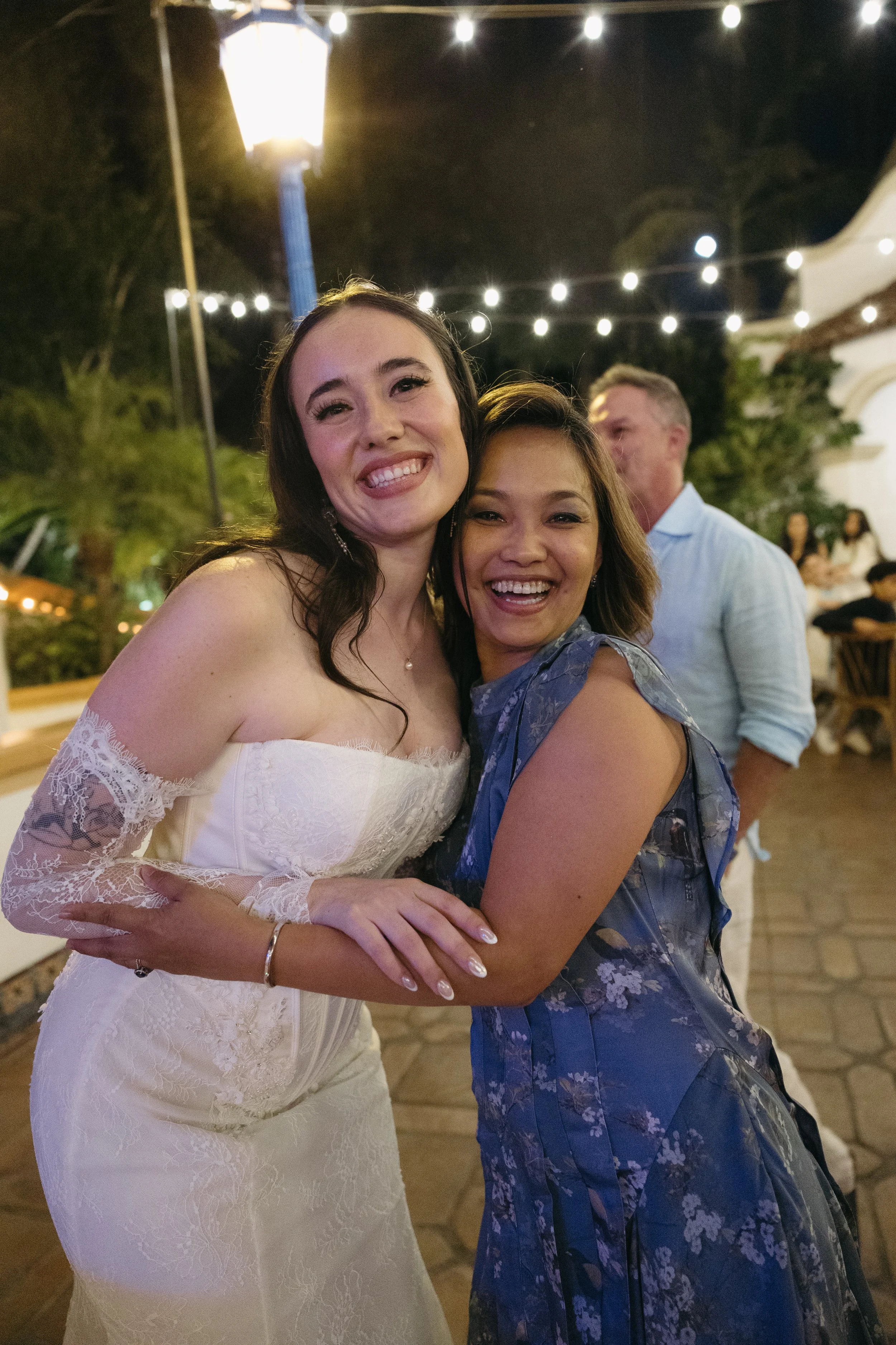 Two women smiling and hugging at an outdoor evening event with string lights overhead.