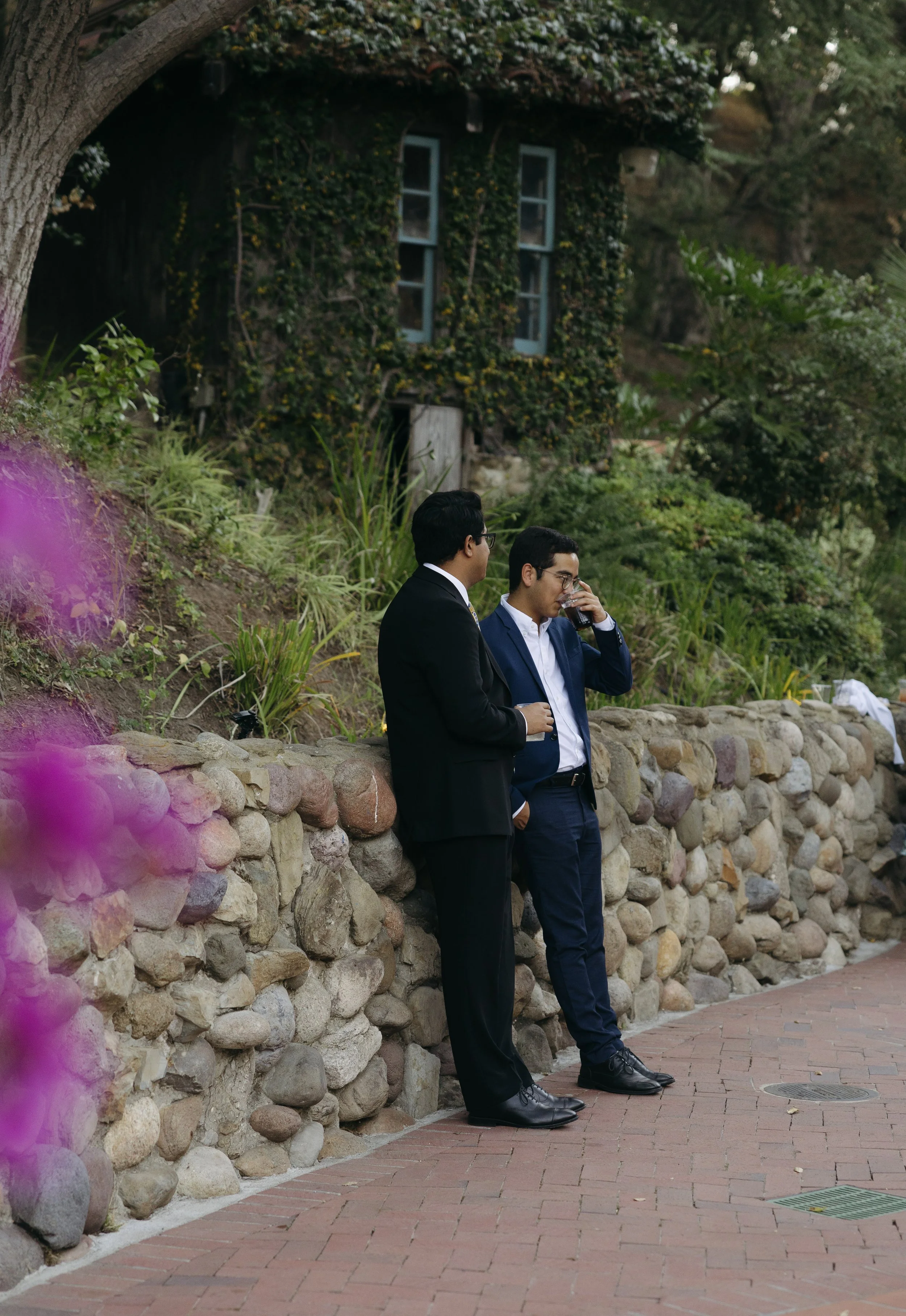 Two men in suits standing and talking against a stone wall, one of them drinking from a glass, with a house covered in ivy and trees in the background.