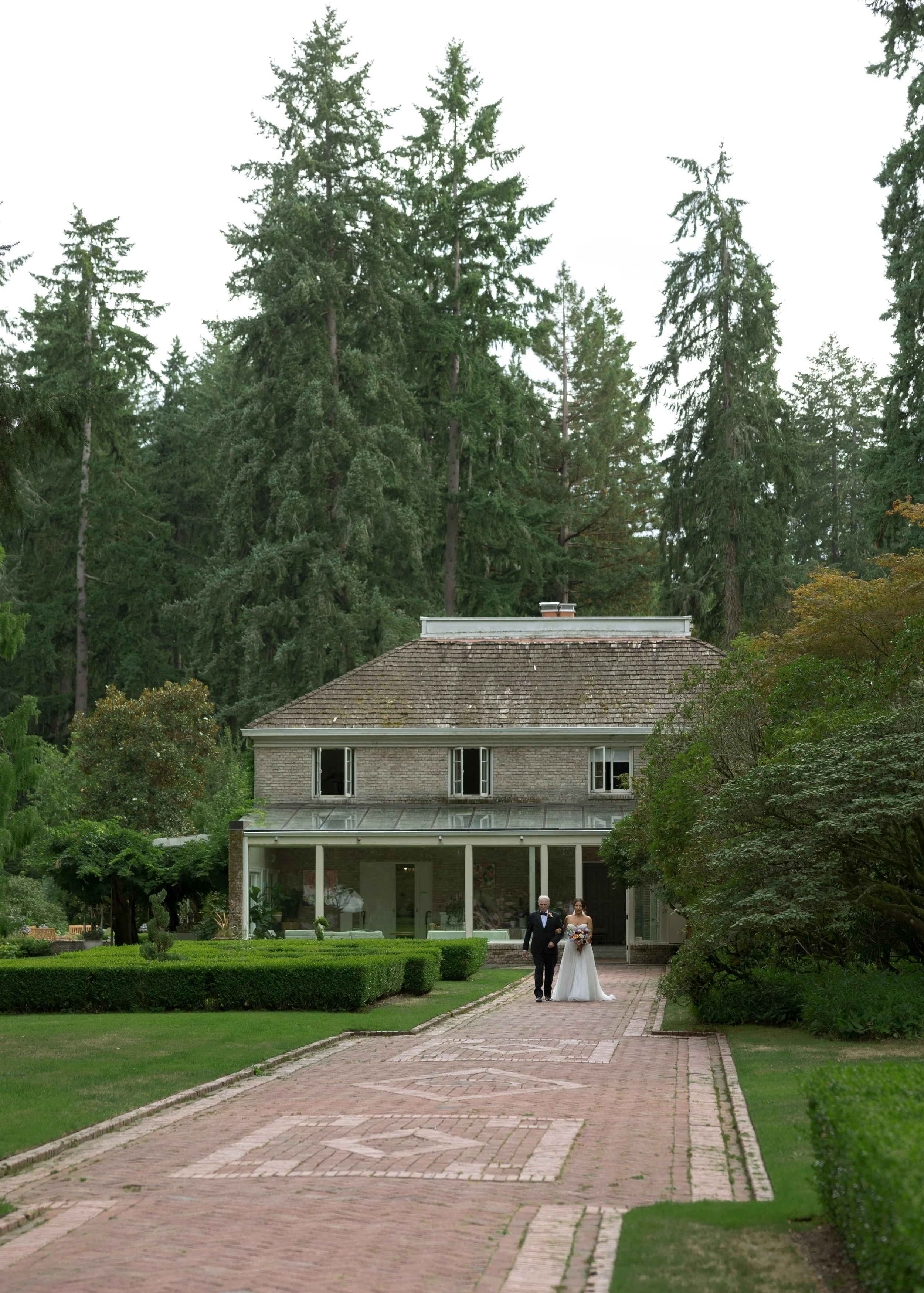 A bride in a white wedding dress walking with her father on a brick pathway leading to a large, historic house surrounded by tall trees and lush greenery at a European inspired Lakewold Gardens wedding in Seattle