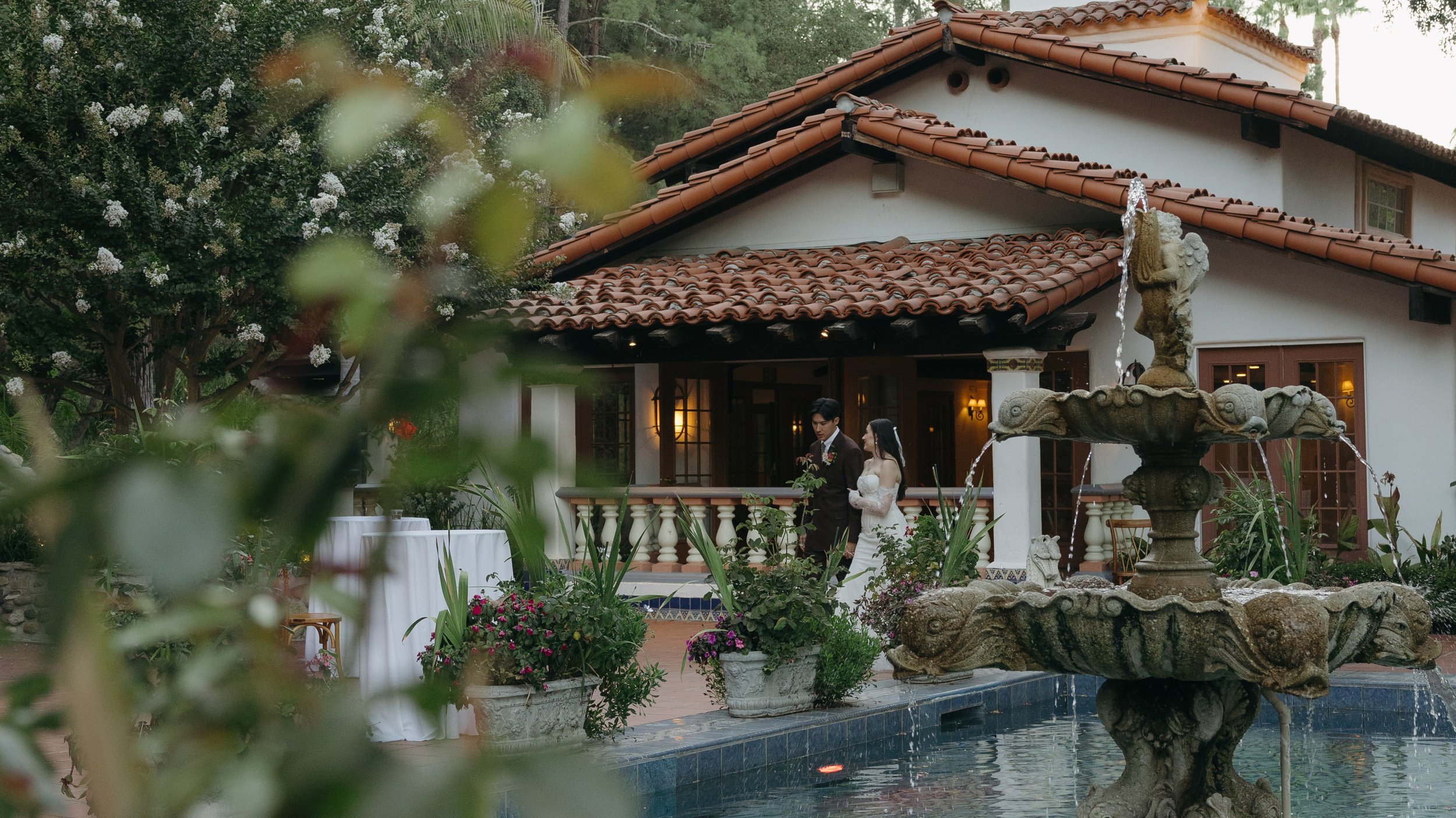 A bride and groom standing together on a porch in front of a house with a tiled roof. In the foreground, there is a fountain with stone sculptures and overflowing water, surrounded by potted plants and greenery at a Rancho Las Lomas Wedding in LA