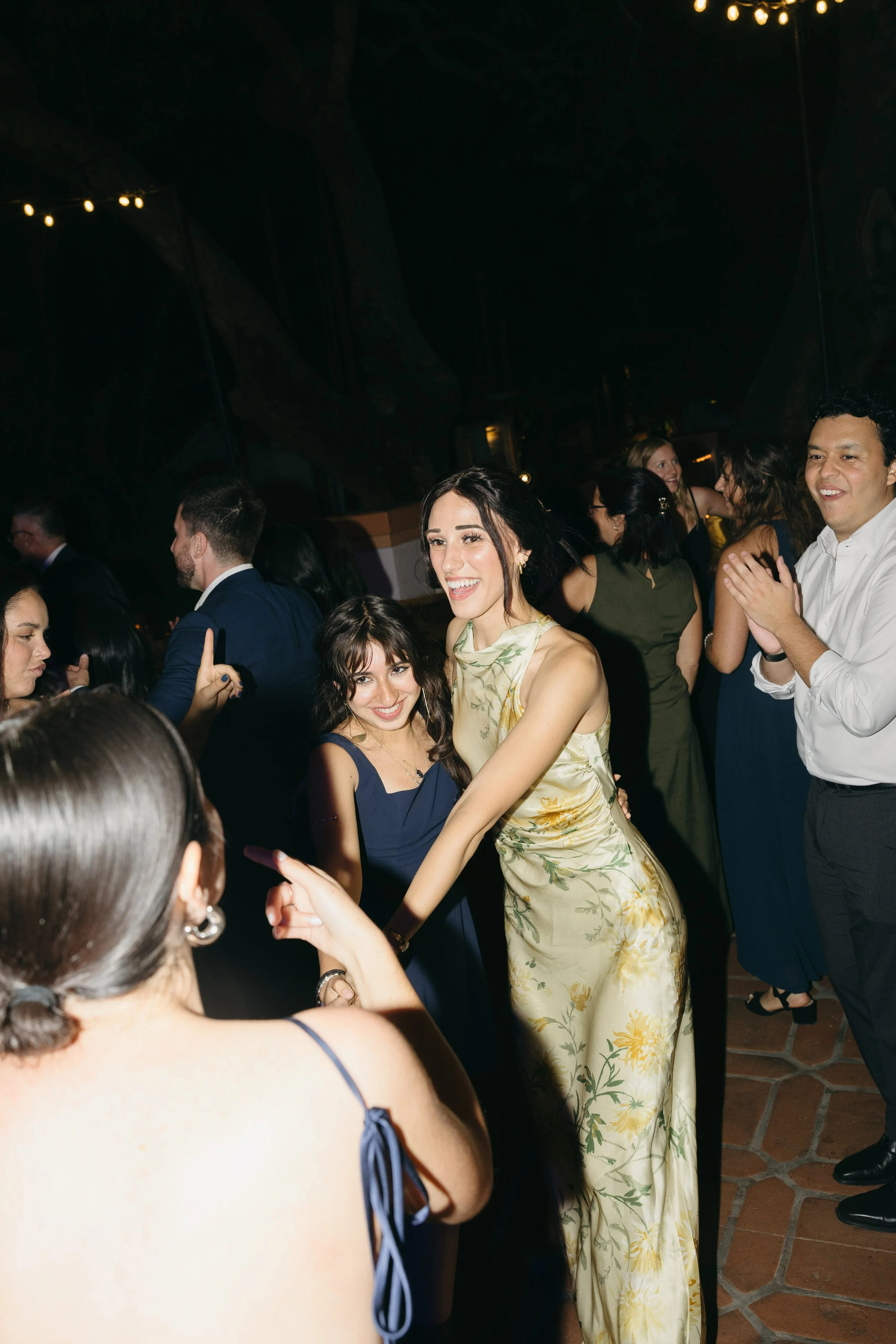 People dancing and celebrating at a party or wedding reception, with string lights overhead and a dark background.
