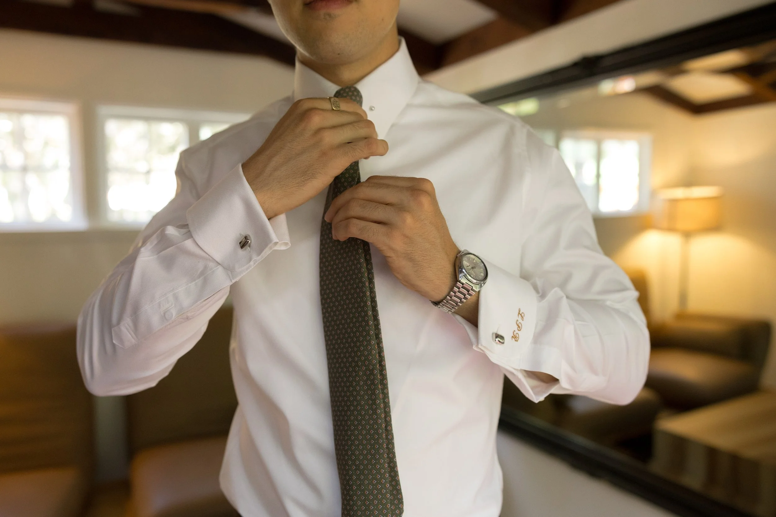 A man in a white dress shirt adjusts his tie, wearing a wristwatch, in a warmly lit room with a mirror, window, and lamp in the background.