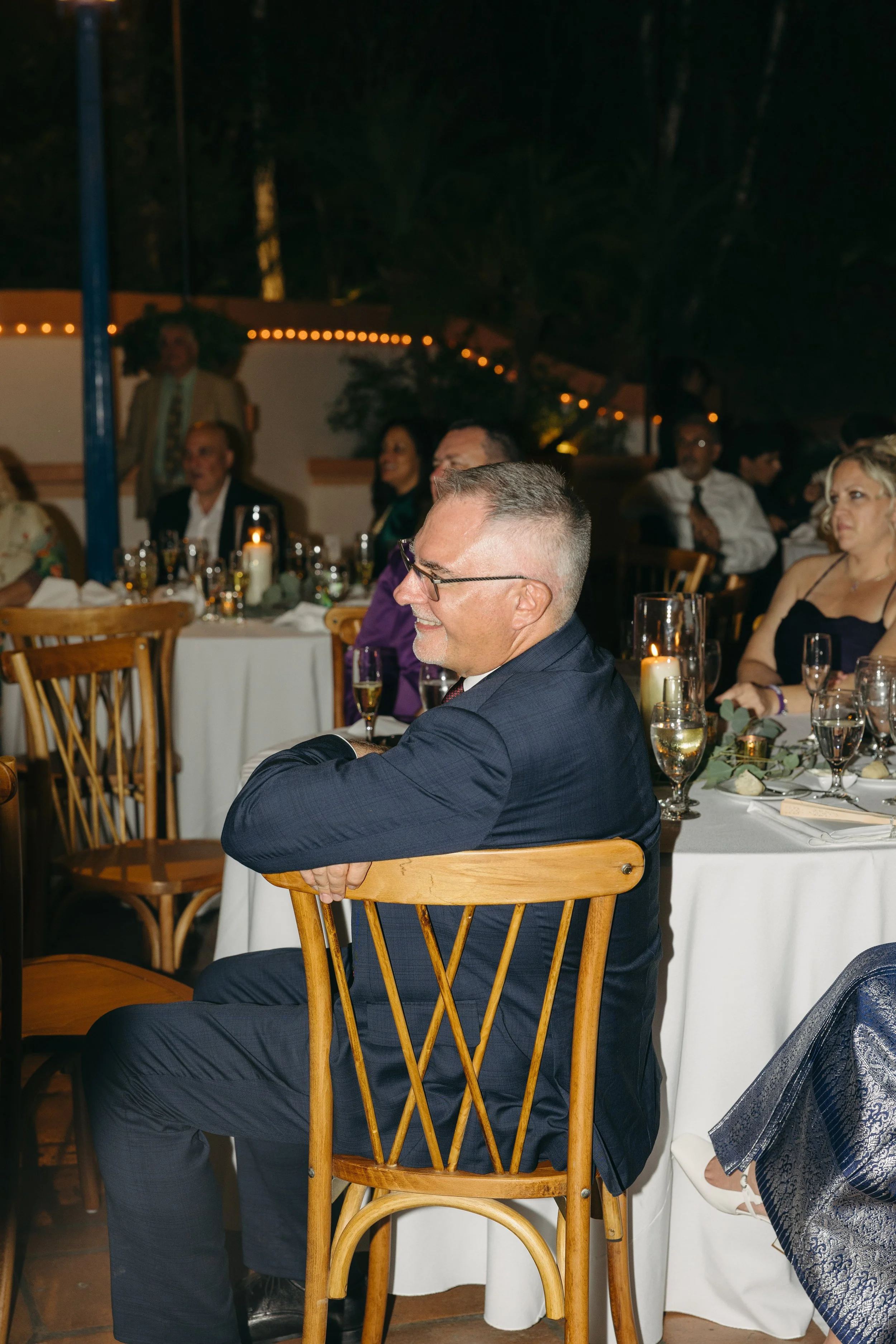 A man in a dark suit, glasses, and a smile sitting at a wooden chair during a formal event or celebration, with decorated tables and other guests in the background.