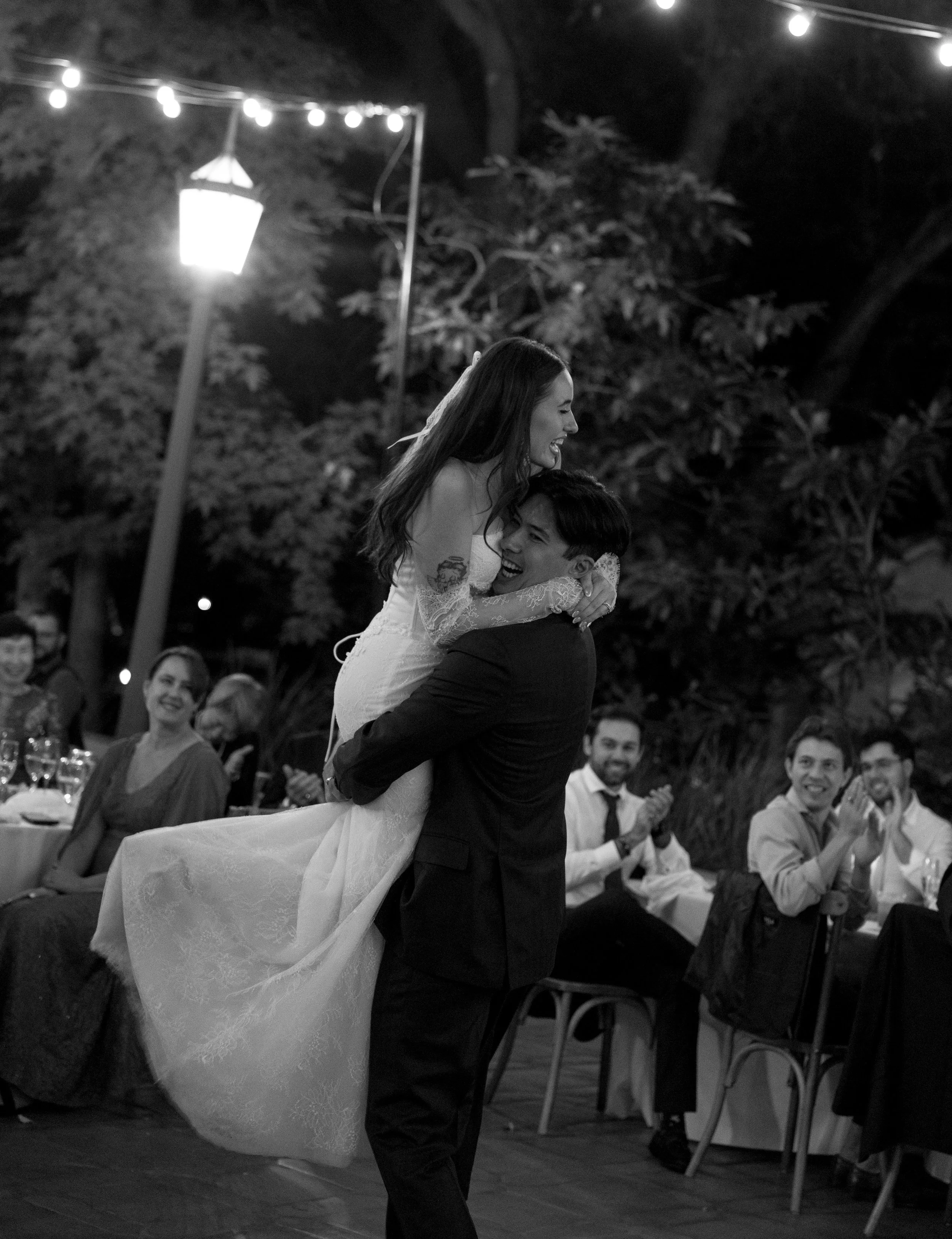 A wedding reception scene in black and white where a groom lifts and dances with the bride. The bride is smiling and looking at the groom, and the guests are sitting at tables in the background, clapping and smiling.