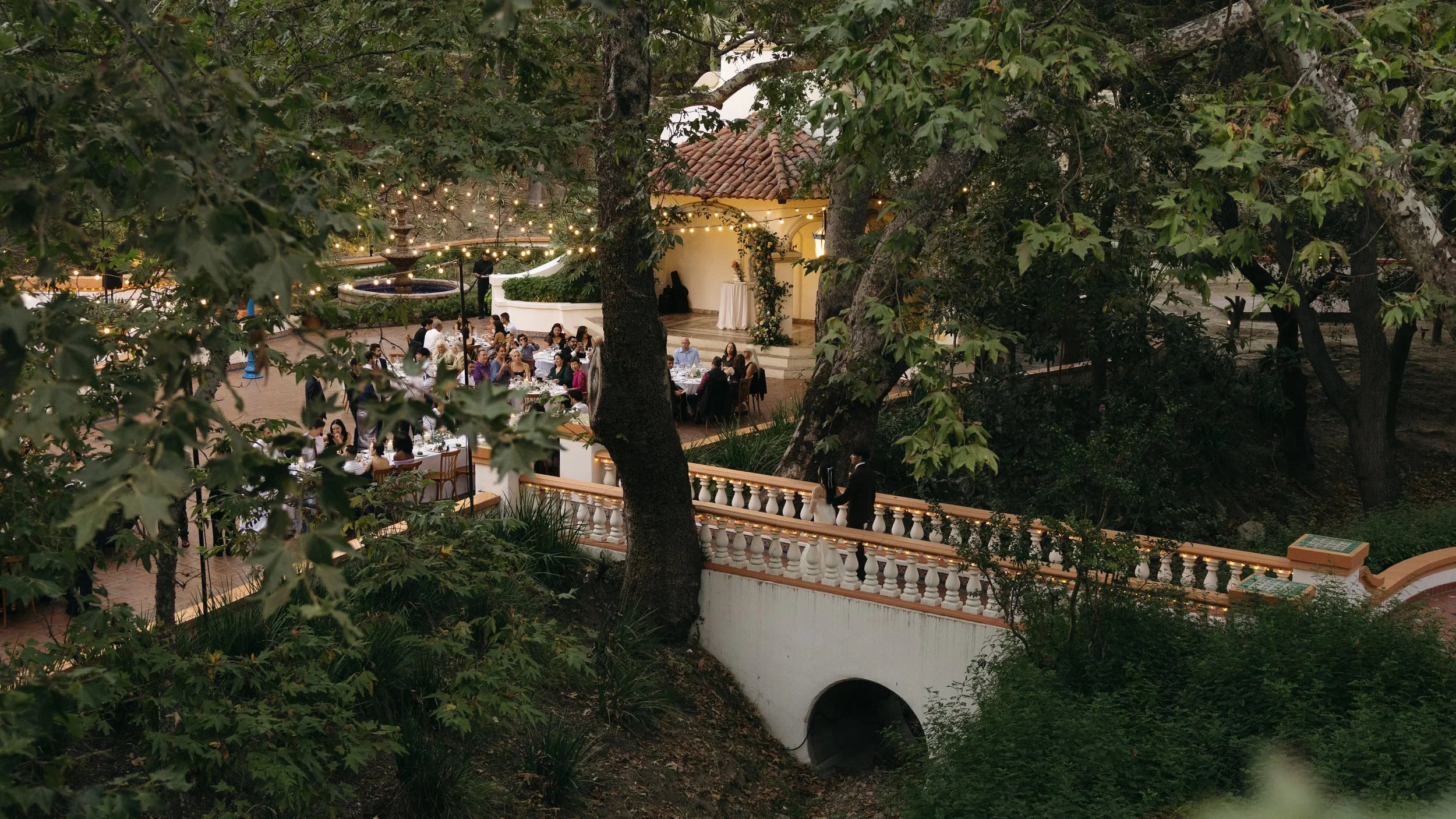 An outdoor wedding reception with guests seated at round tables, string lights hung overhead, and a couple walking on a decorative bridge surrounded by trees and greenery at a Rancho Las Lomas Wedding in Los Angeles