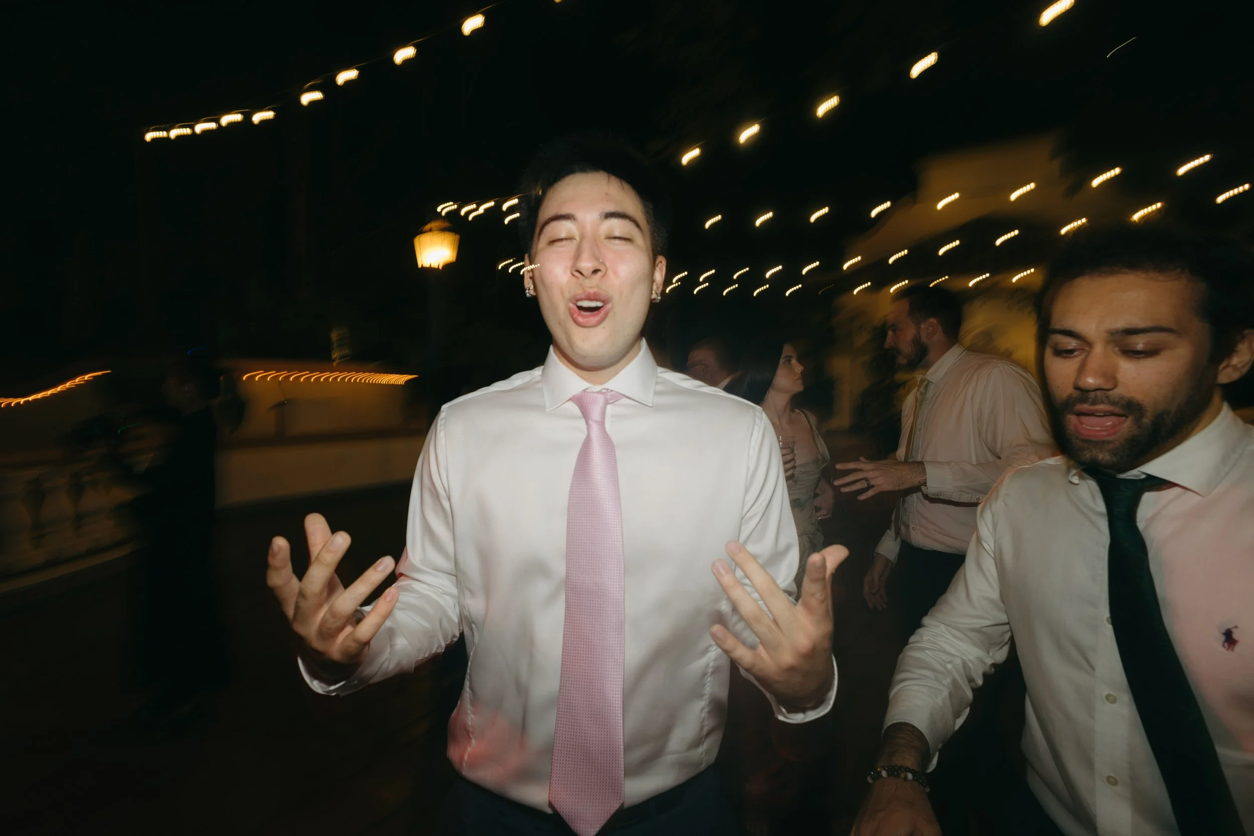 Young man in a white shirt, pink tie, and earrings dancing and singing at a party or celebration at night, with other guests in the background.
