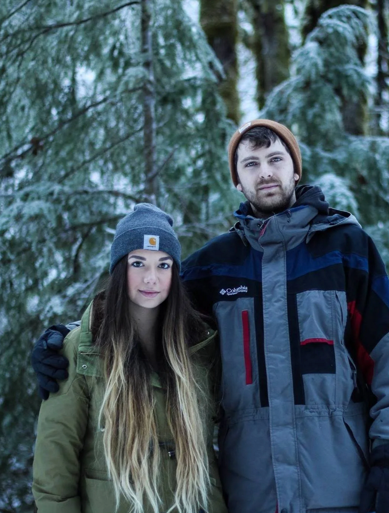 A young man and woman stand together outdoors in a snowy forest, dressed in winter clothing. The woman has long brown hair, a gray beanie, and an olive green jacket. The man has short dark hair, a brown beanie, and a gray and black Columbia jacket.