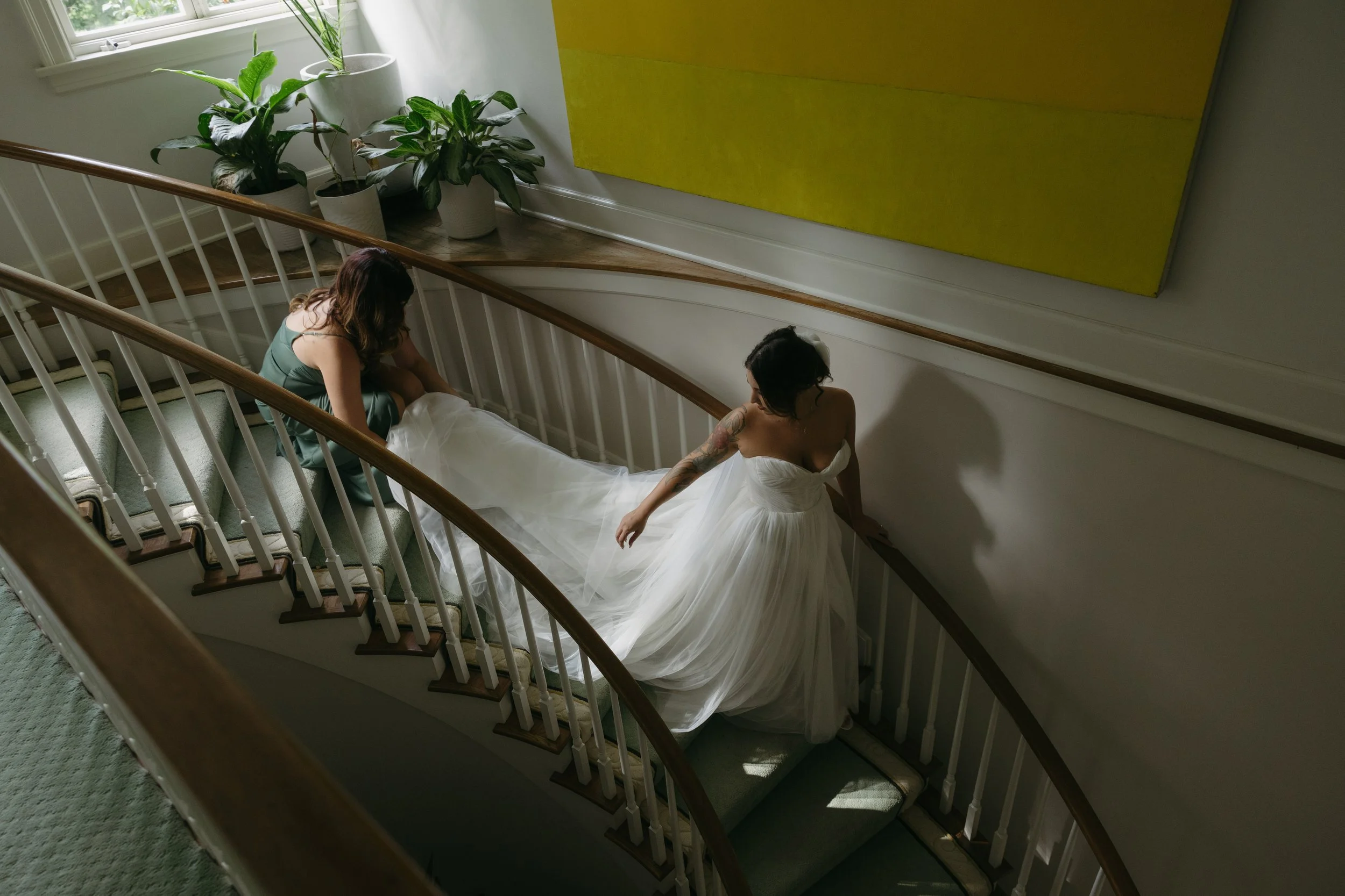 A bride in a white wedding gown walking down a curved staircase with a woman in a green dress adjusting her dress.