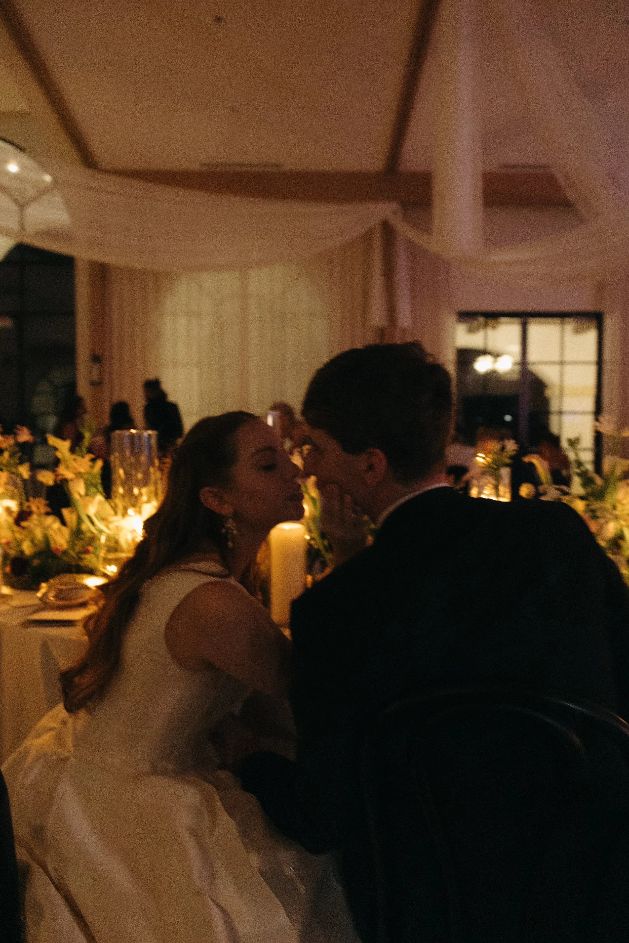 A bride and groom sharing a kiss at their wedding reception, warmly lit and decorated with flowers and candles.