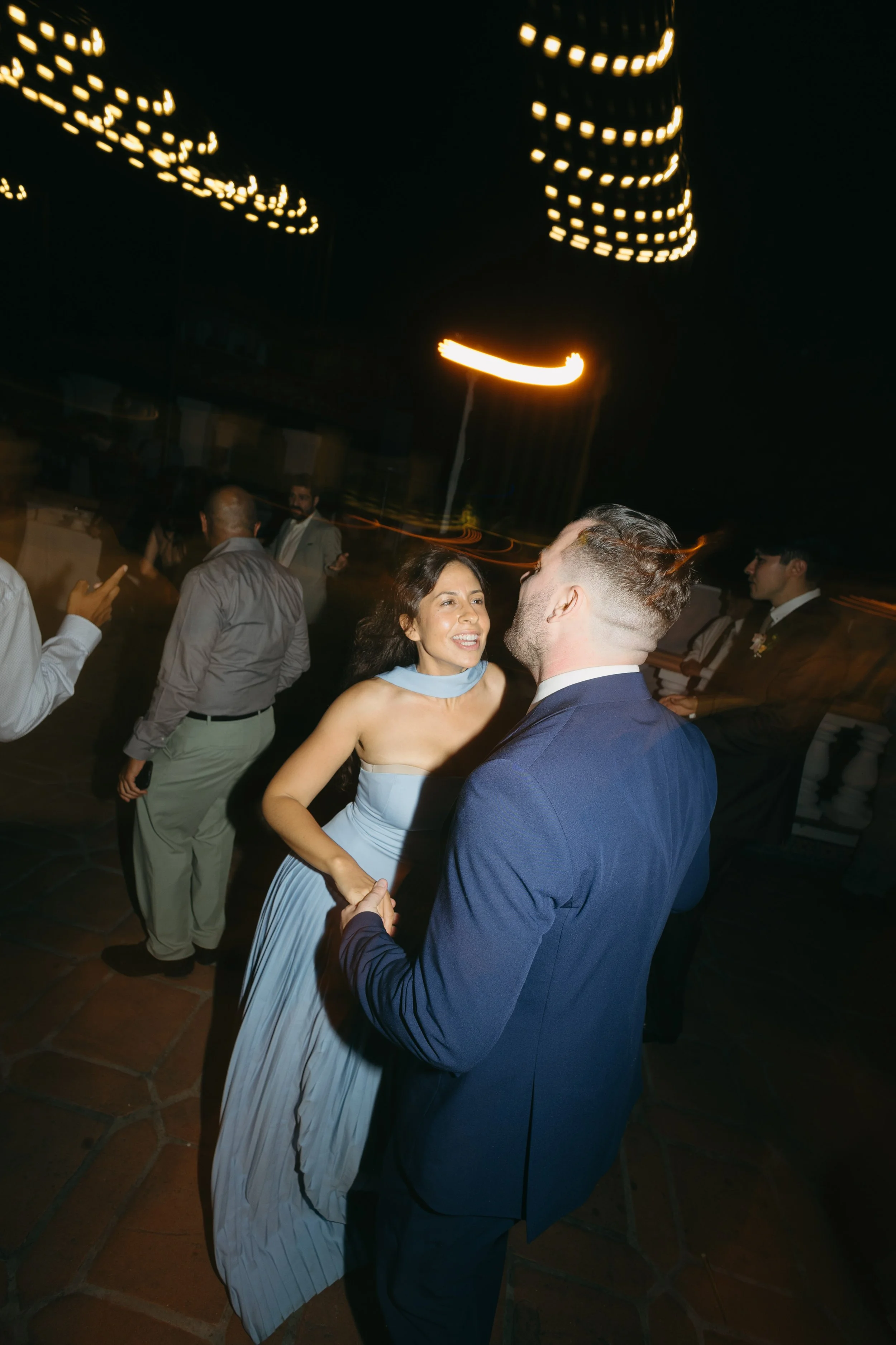A woman in a light blue dress and a man in a dark blue suit are dancing closely on a dance floor at night, with a blurred background of other people and city lights.