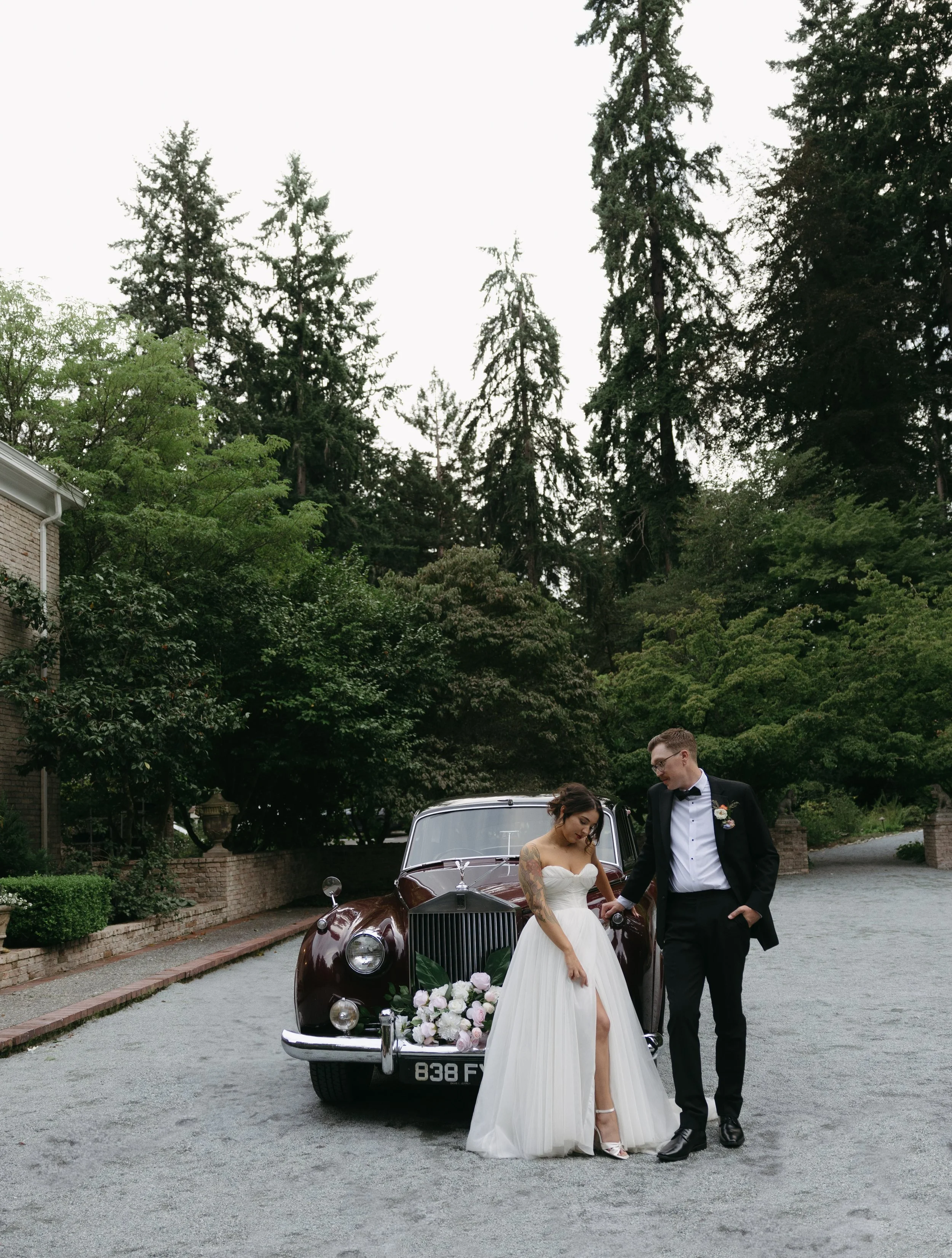Bride and groom standing next to a vintage car decorated with flowers outdoors, with tall trees and greenery in the background.