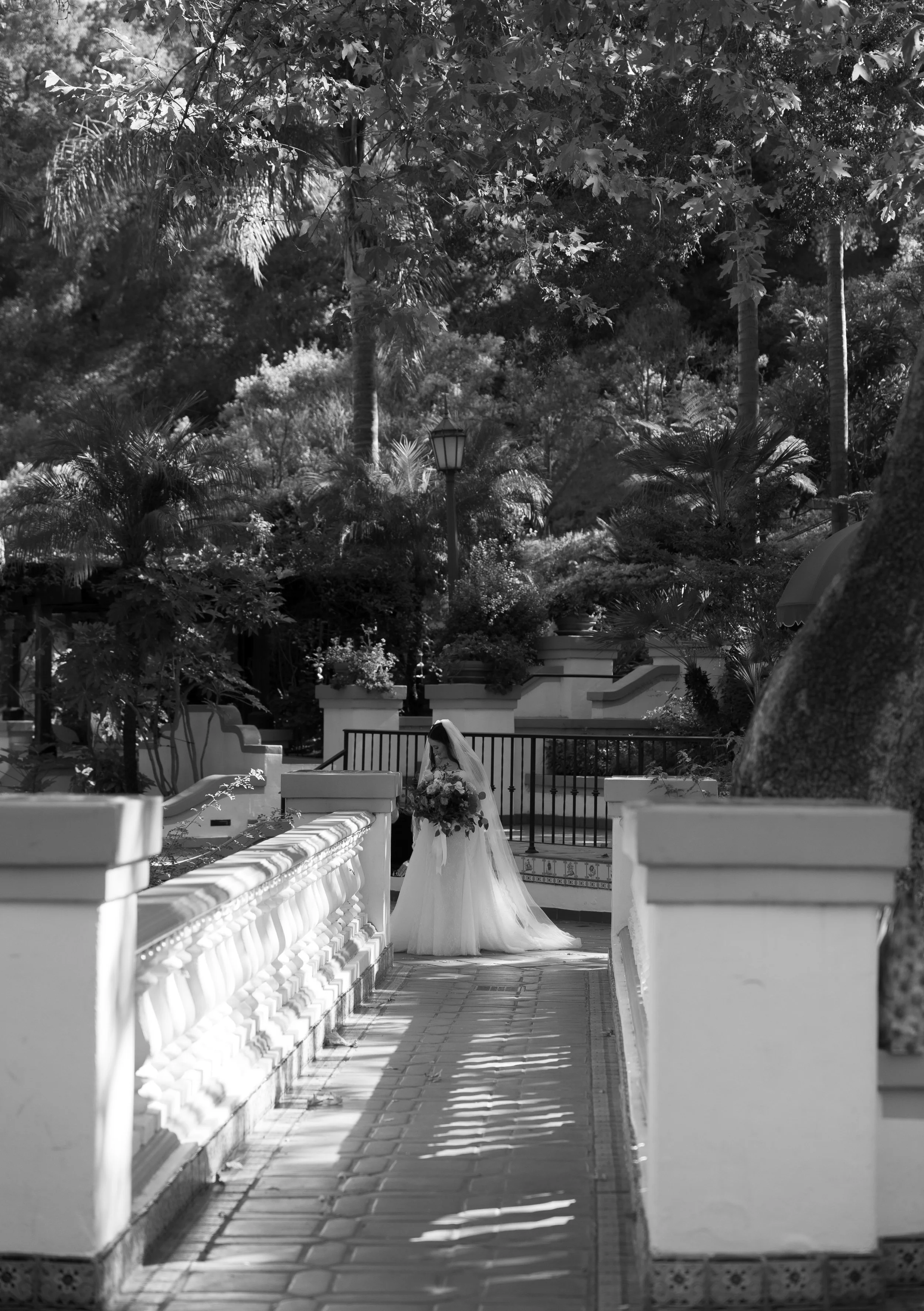 A bride in a wedding dress holding a bouquet of flowers in a garden with lush trees and plants, sunlight casting shadows on a paved walkway at a Rancho Las Lomas Wedding in Los Angeles