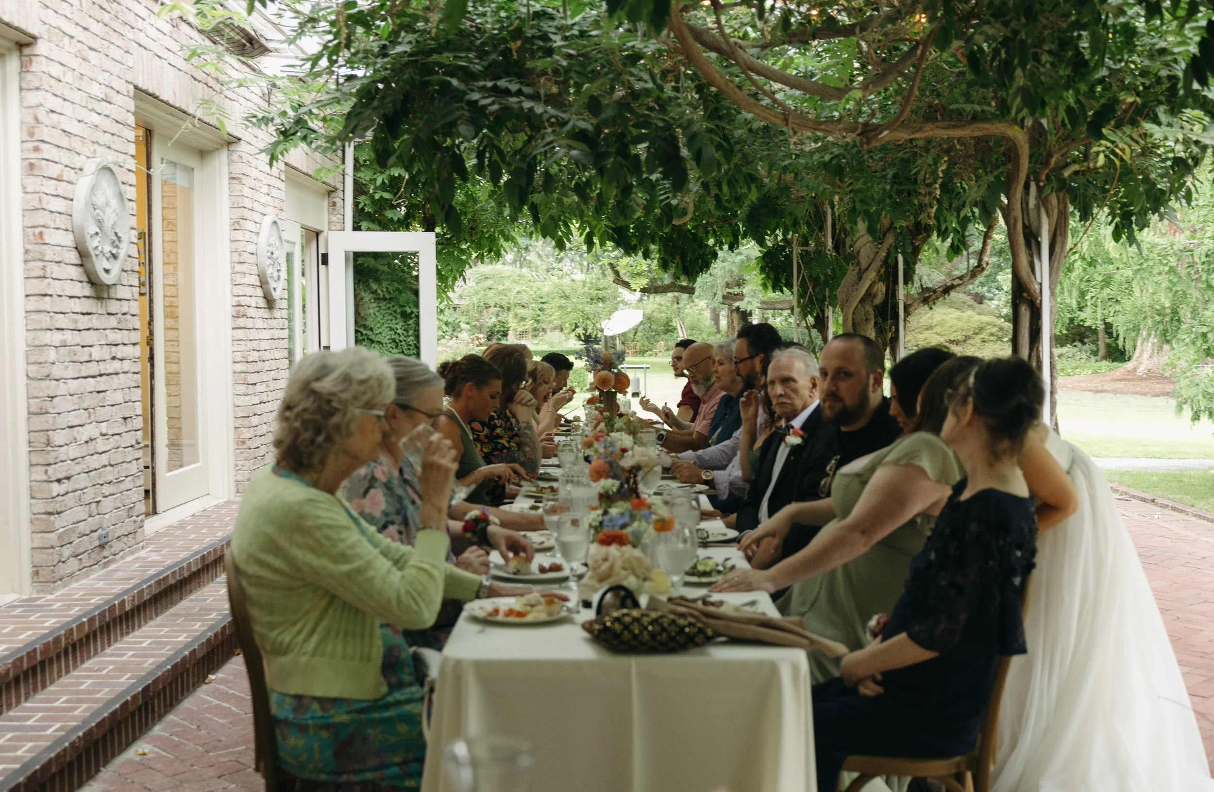 An outdoor dinner wedding reception, with guests seated at a long dining table outdoors under a tree, enjoying a meal at a European inspired Lakewold Gardens wedding in Seattle