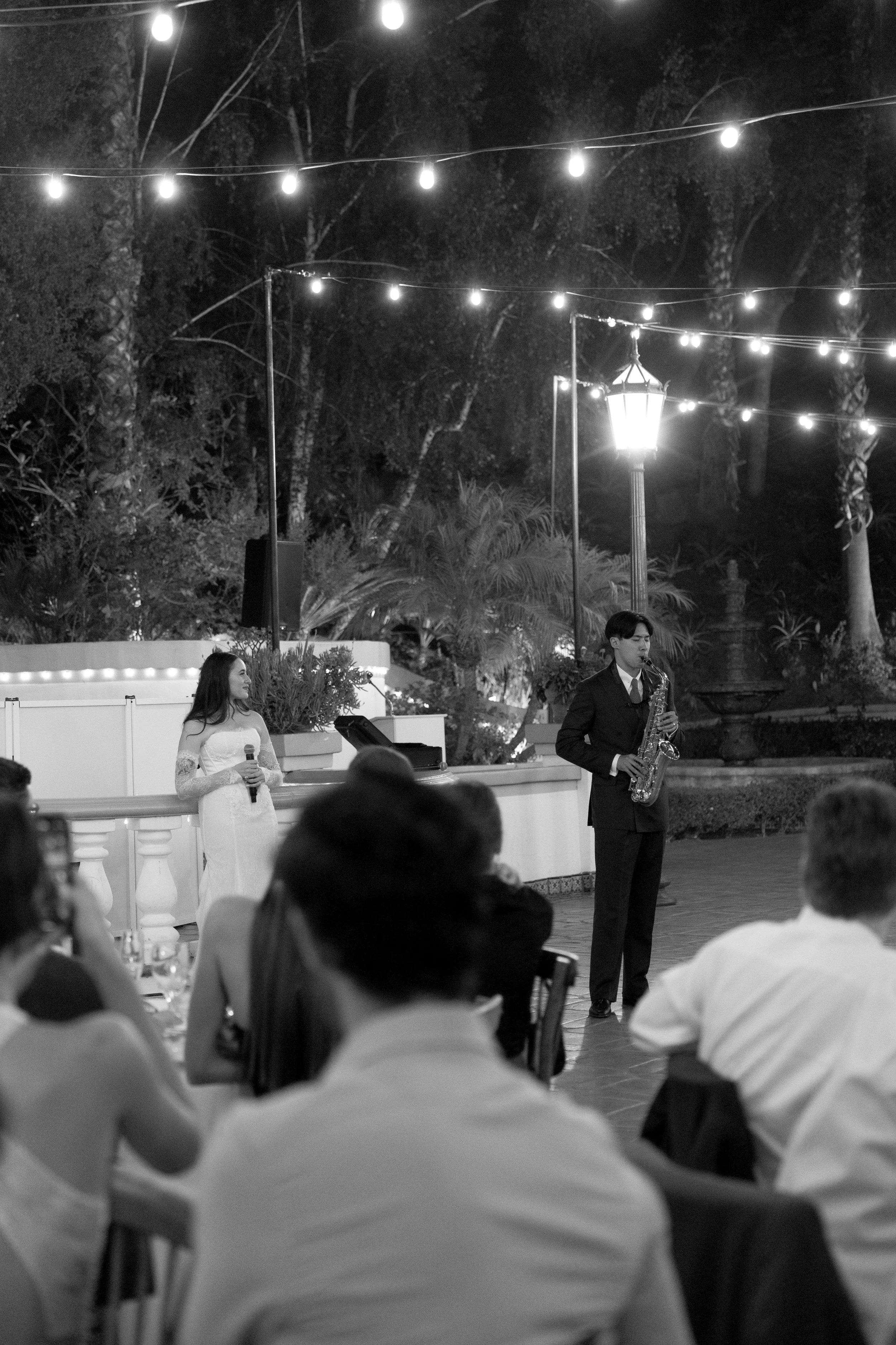 An outdoor nighttime event with string lights, a band with a female singer holding a microphone, and a male saxophonist performing, while guests in formal attire sit and watch.