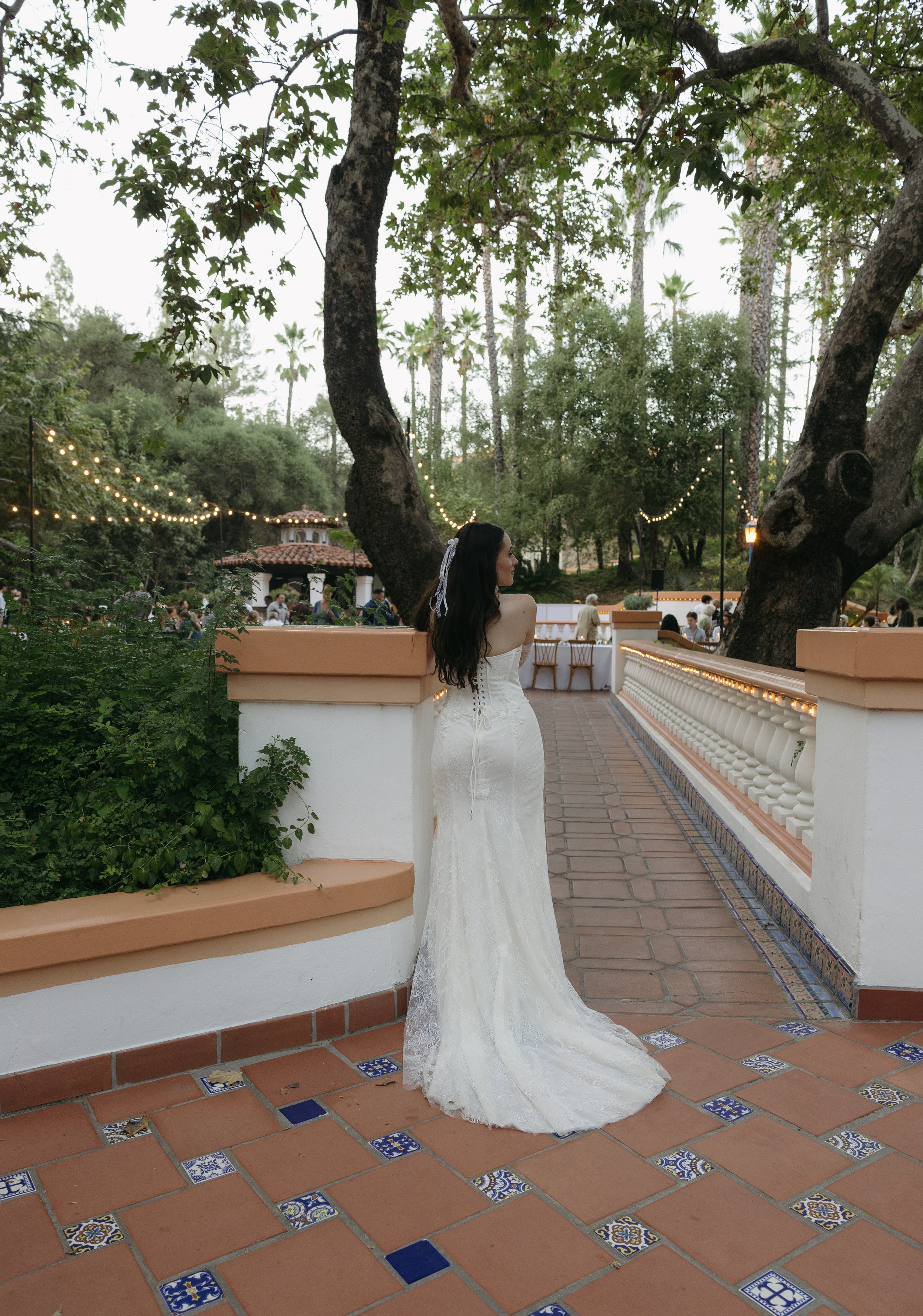 A woman in a white wedding dress with dark hair, standing on a terracotta tile patio near a white stucco wall with beige trim, in an outdoor garden setting decorated with string lights and large trees, at sunset or early evening.