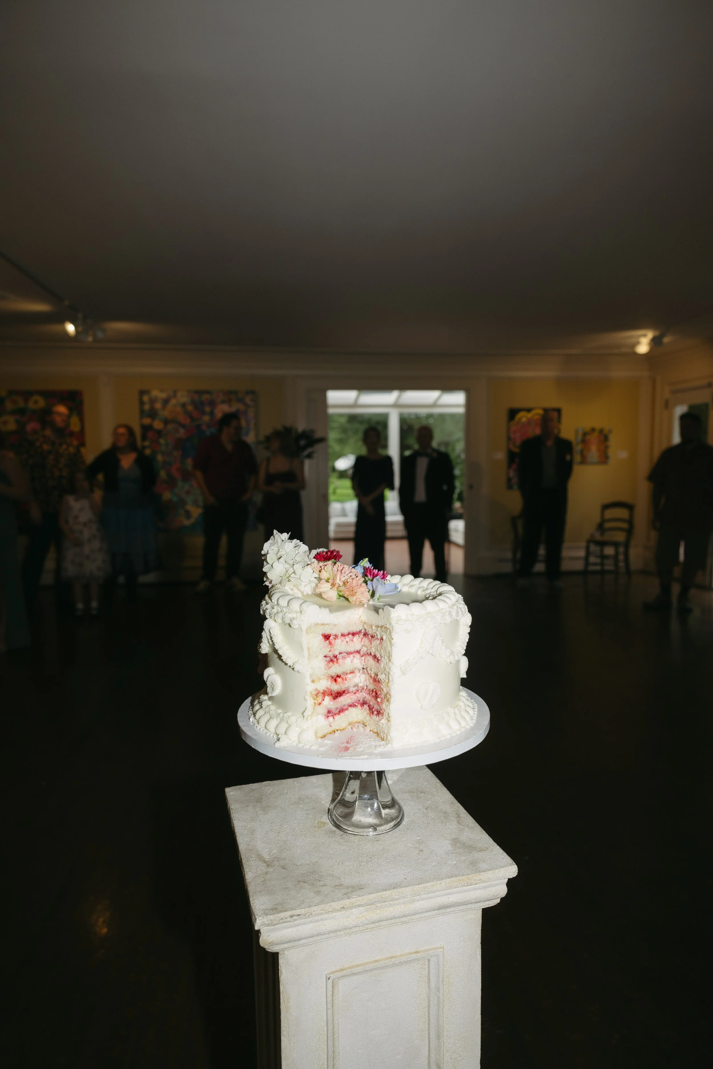 A white wedding cake with pink and white flowers on top, partially cut to show red and white layers, placed on a cake stand on a pedestal in a dimly lit room with people in the background.