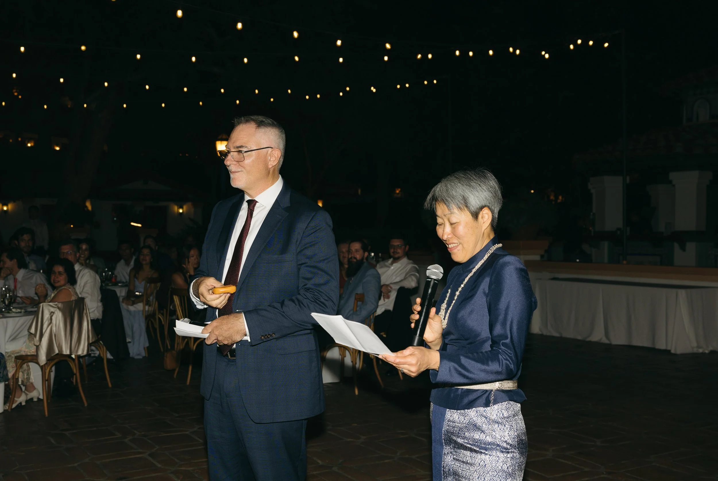 A woman in a blue satin dress reading from a paper and speaking into a microphone, standing next to a man in a blue suit at an evening event with string lights overhead.