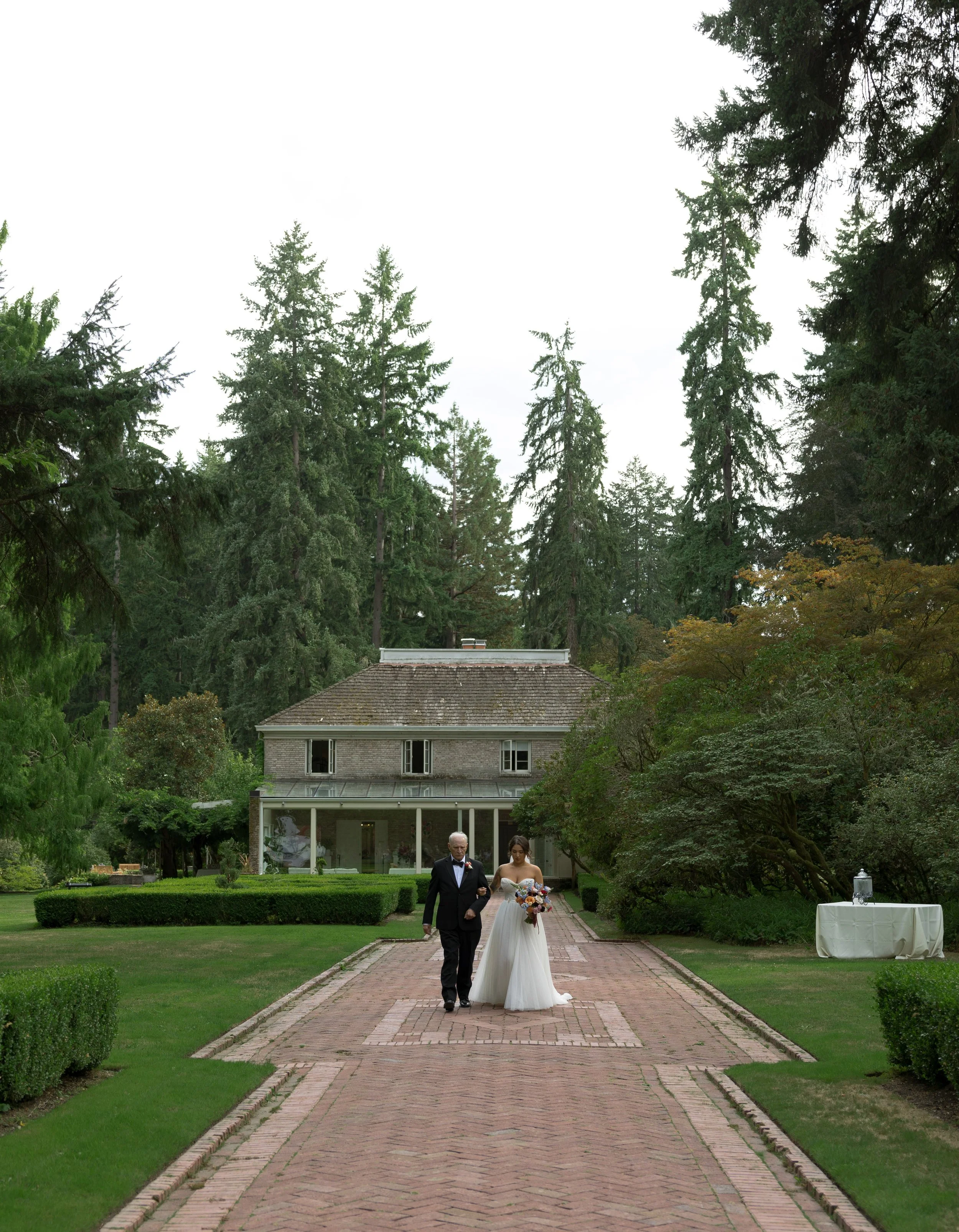A bride in a white wedding dress being escorted down a brick pathway by her father, at a European inspired Lakewold Gardens wedding in Seattle