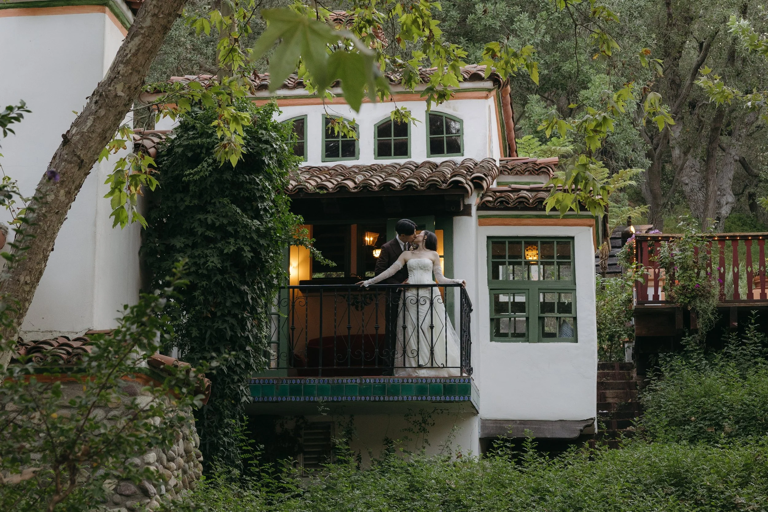A bride and groom sharing a kiss on a balcony of a white house with green window frames, surrounded by trees and greenery at a Rancho Las Lomas Wedding in Los Angeles